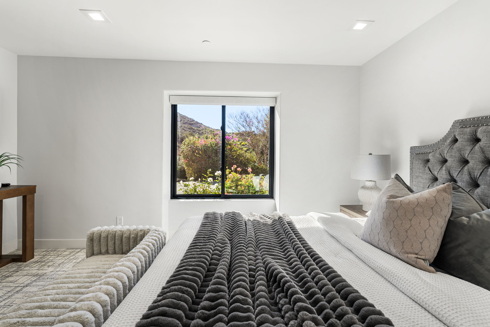 This primary bedroom features a plush, tufted gray headboard and a textured charcoal throw blanket, creating a cozy and sophisticated atmosphere. A large window serves as a focal point, flooding the room with natural light and offering a scenic view of the lush, sunlit landscape outside. The space is complemented by neutral wall tones, minimalist recessed lighting, and a soft-textured bench at the foot of the bed, enhancing the overall sense of modern luxury.