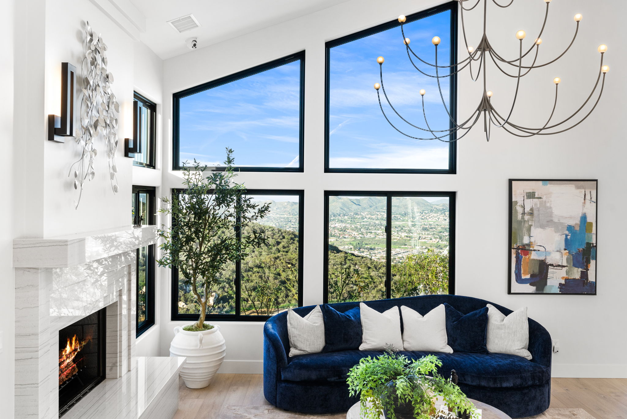 This bright, contemporary living room features a striking blue curved sofa and a sleek marble-clad fireplace. Large, multi-pane windows span the wall, offering breathtaking panoramic views of the surrounding hills and valley. The space is accented by a sculptural modern chandelier and elegant wall decor, creating a sophisticated and luxurious atmosphere.