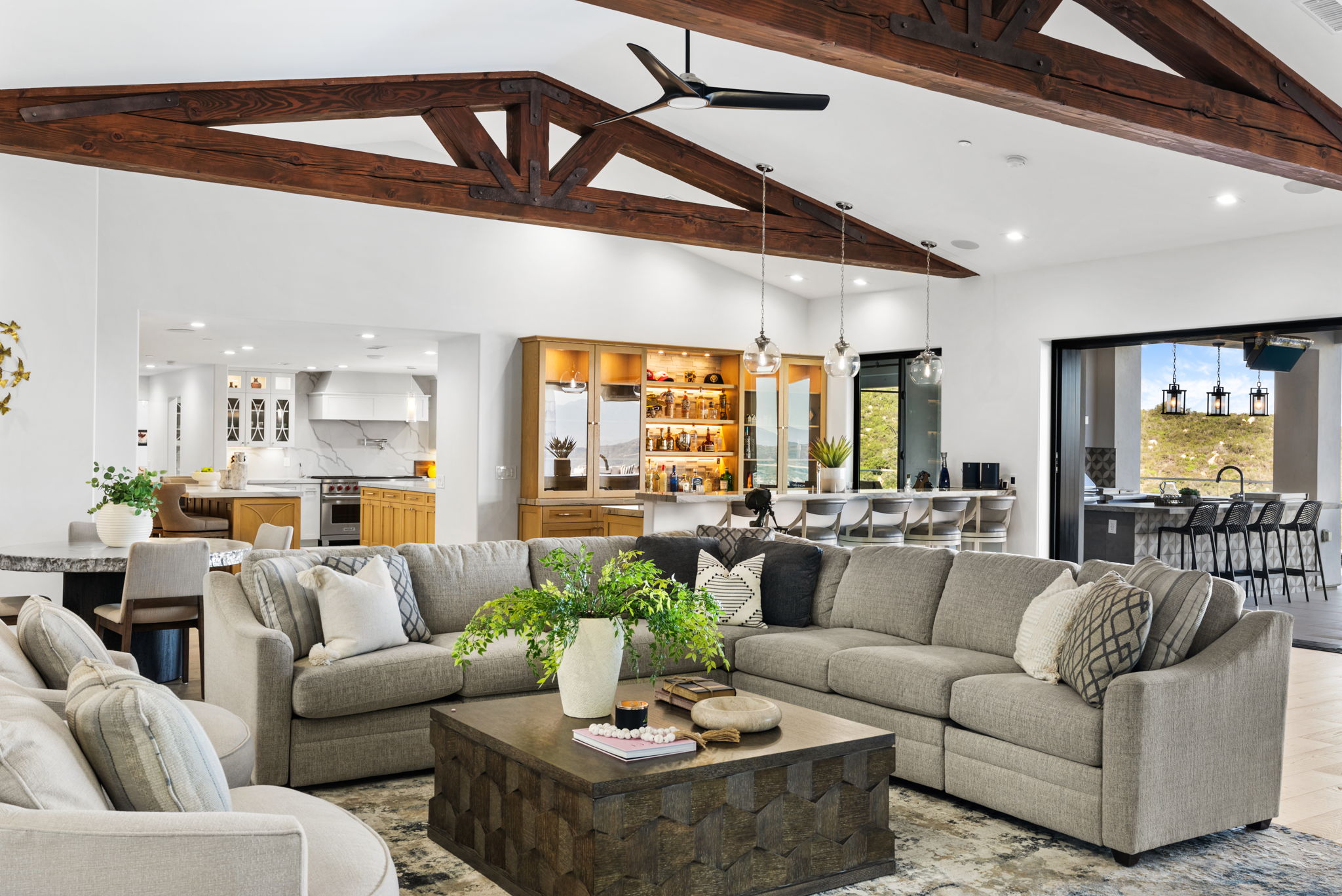 This spacious, open-concept living area features a grand vaulted ceiling with rustic exposed timber beams and a large gray sectional sofa centered around a textured wooden coffee table. The space flows seamlessly into a bar area and a gourmet kitchen, showcasing an elegant blend of modern luxury and farmhouse warmth. Large glass sliding doors offer a glimpse of the outdoor patio, enhancing the bright and airy atmosphere perfect for entertaining.