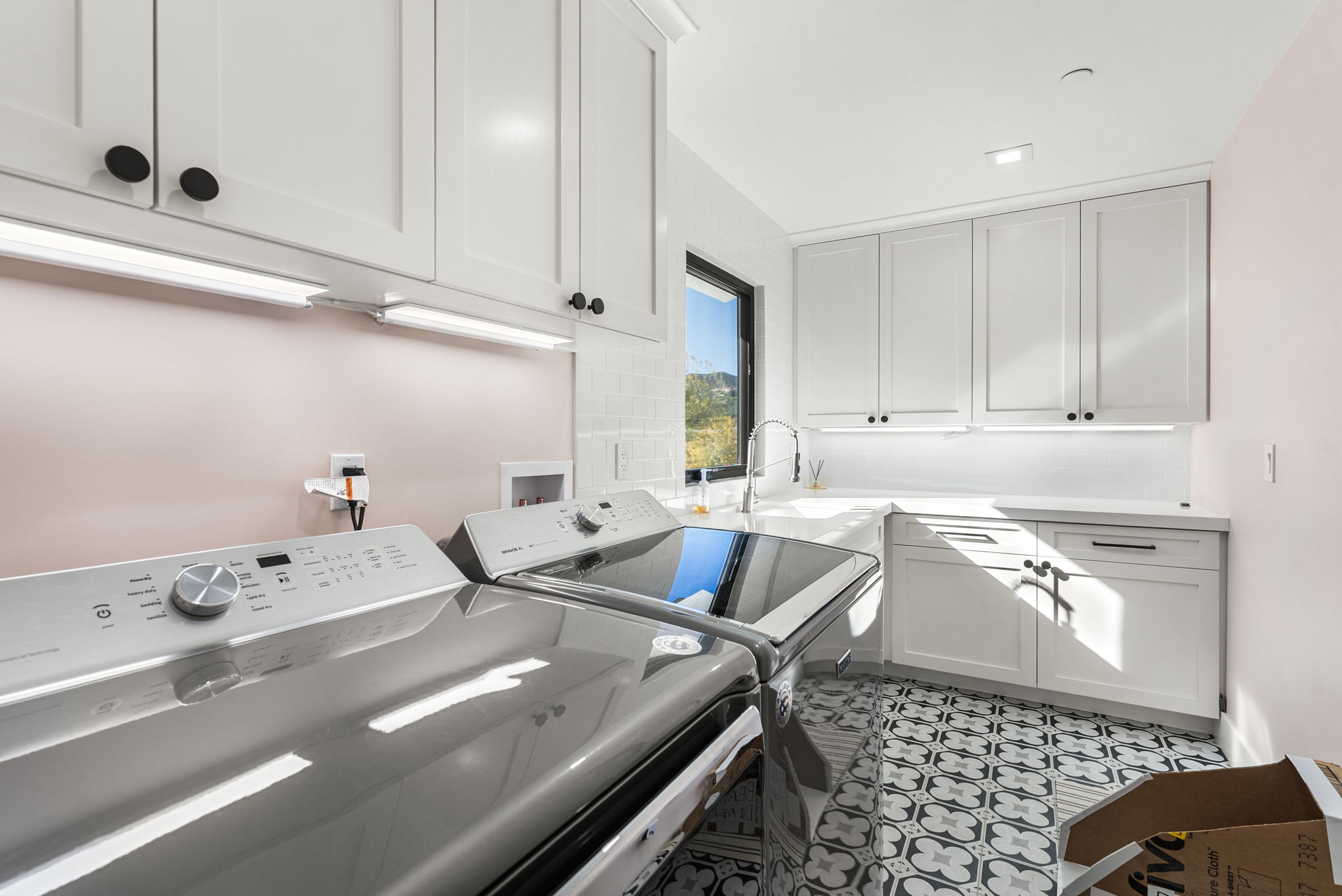 This bright and functional laundry room features sleek white cabinetry, a countertop workstation with an integrated sink, and modern gray patterned floor tiles. The space is equipped with metallic, high-efficiency washer and dryer units, complemented by under-cabinet task lighting and a soft pink wall accent. It presents a clean, contemporary aesthetic with ample storage, making it a highly practical and stylish utility space.