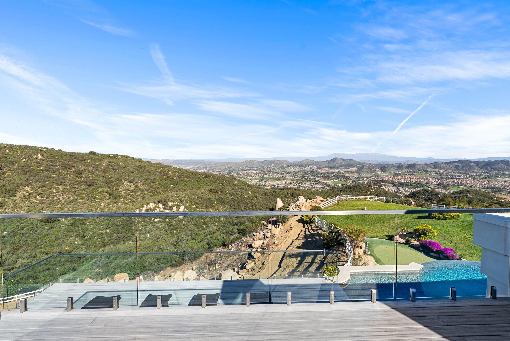 This stunning elevated deck offers breathtaking panoramic views of the surrounding valley and mountains. The space features a modern glass railing, a sleek wood-plank deck surface, and direct access to an infinity-edge pool, creating a seamless blend of luxury and nature. It is an ideal outdoor retreat for relaxation and entertainment with its expansive outlook.