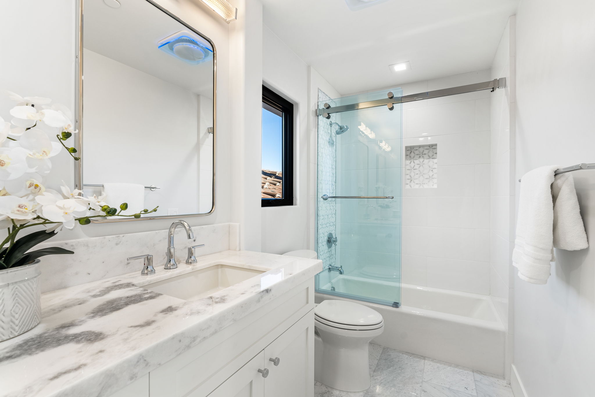 This clean, modern bathroom features a white marble vanity countertop with polished chrome fixtures and a sleek, frameless glass sliding door over the shower/tub combination. The space is accented by patterned tile inlay in the shower niche and a bright, minimalist aesthetic. A window allows natural light to flow into the room, creating an inviting and well-maintained feel.