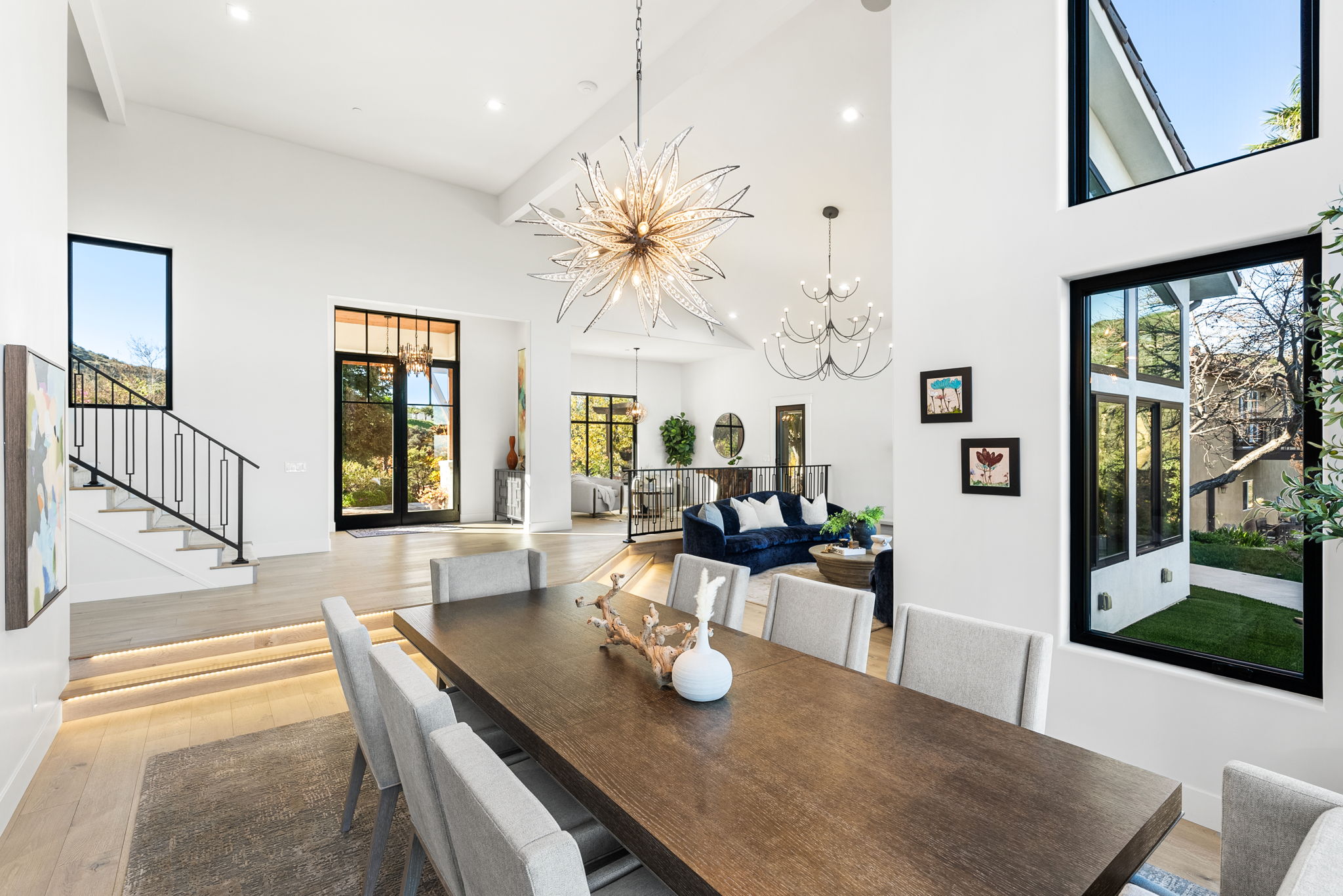 This elegant open-concept dining room features a large dark wood table surrounded by plush grey chairs, illuminated by a striking starburst chandelier. The space seamlessly transitions into a sunken living area with a deep blue velvet sofa and high ceilings, emphasizing an airy, luxurious atmosphere. Modern architectural details, including stair lighting and floor-to-ceiling windows, highlight the sophisticated design and expansive layout of the home.