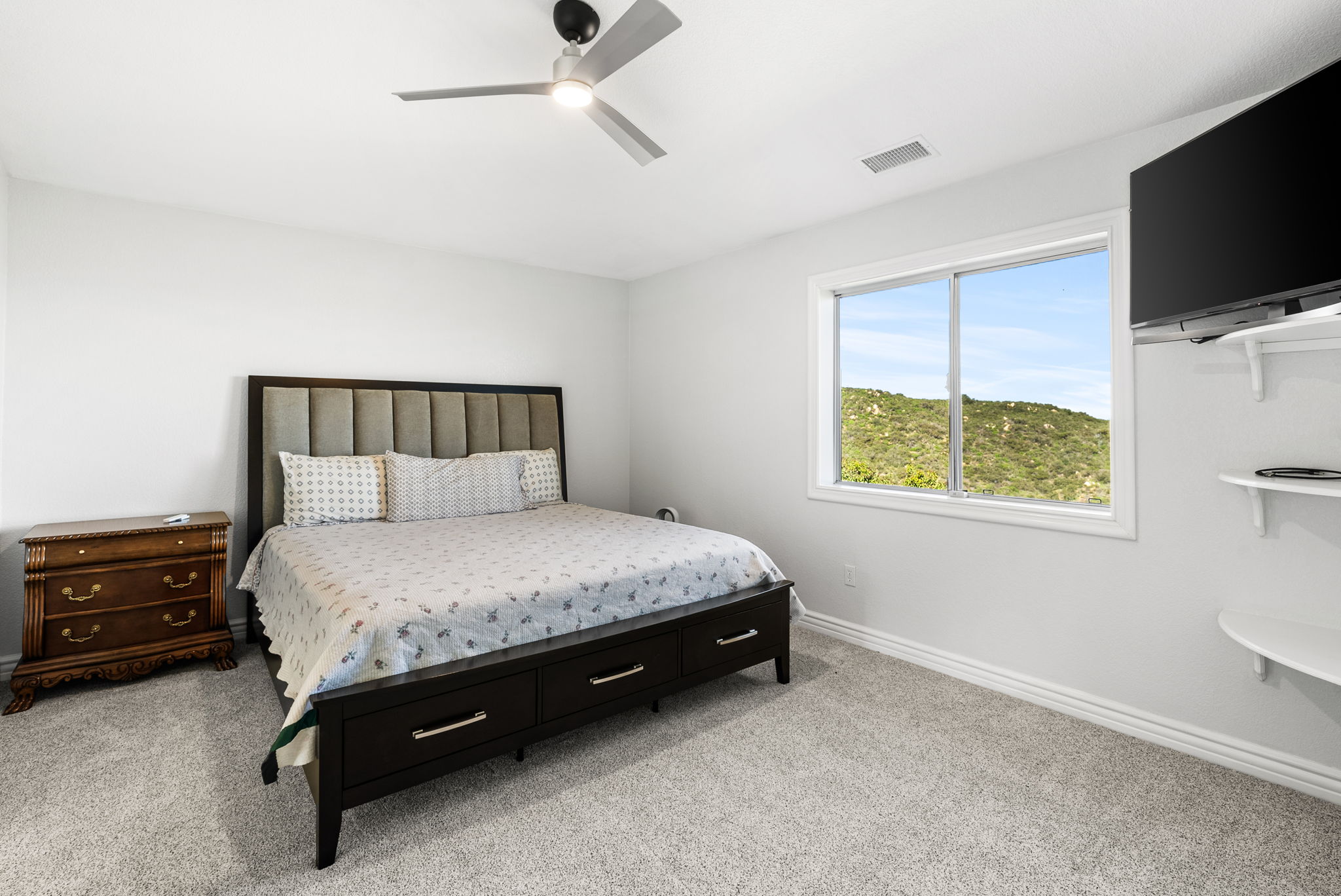 This guest bedroom features a dark wood bed frame with integrated storage drawers and a neutral upholstered headboard. The room includes a classic wooden nightstand, a ceiling fan, and a flat-screen TV wall-mounted on floating shelves, all set against a clean white wall backdrop with carpeted flooring. Large windows provide natural light and a pleasant view of the surrounding hilly landscape.