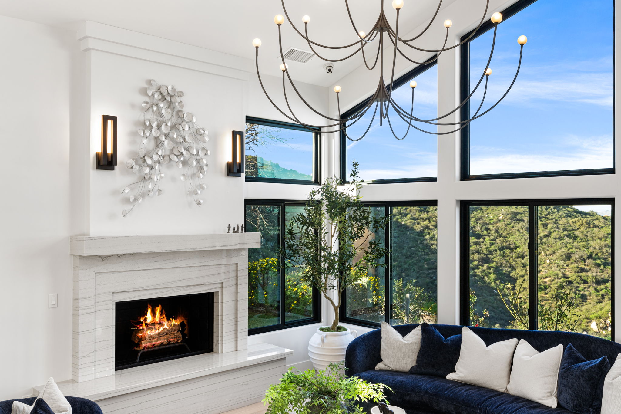 This elegant living room features a striking stone fireplace accented by modern wall sconces and a unique metal wall art installation. A large, navy blue velvet sofa sits adjacent to floor-to-ceiling windows that provide abundant natural light and breathtaking views of the surrounding hillside landscape. The space is crowned by a contemporary, sputnik-style chandelier, creating a sophisticated and inviting atmosphere.