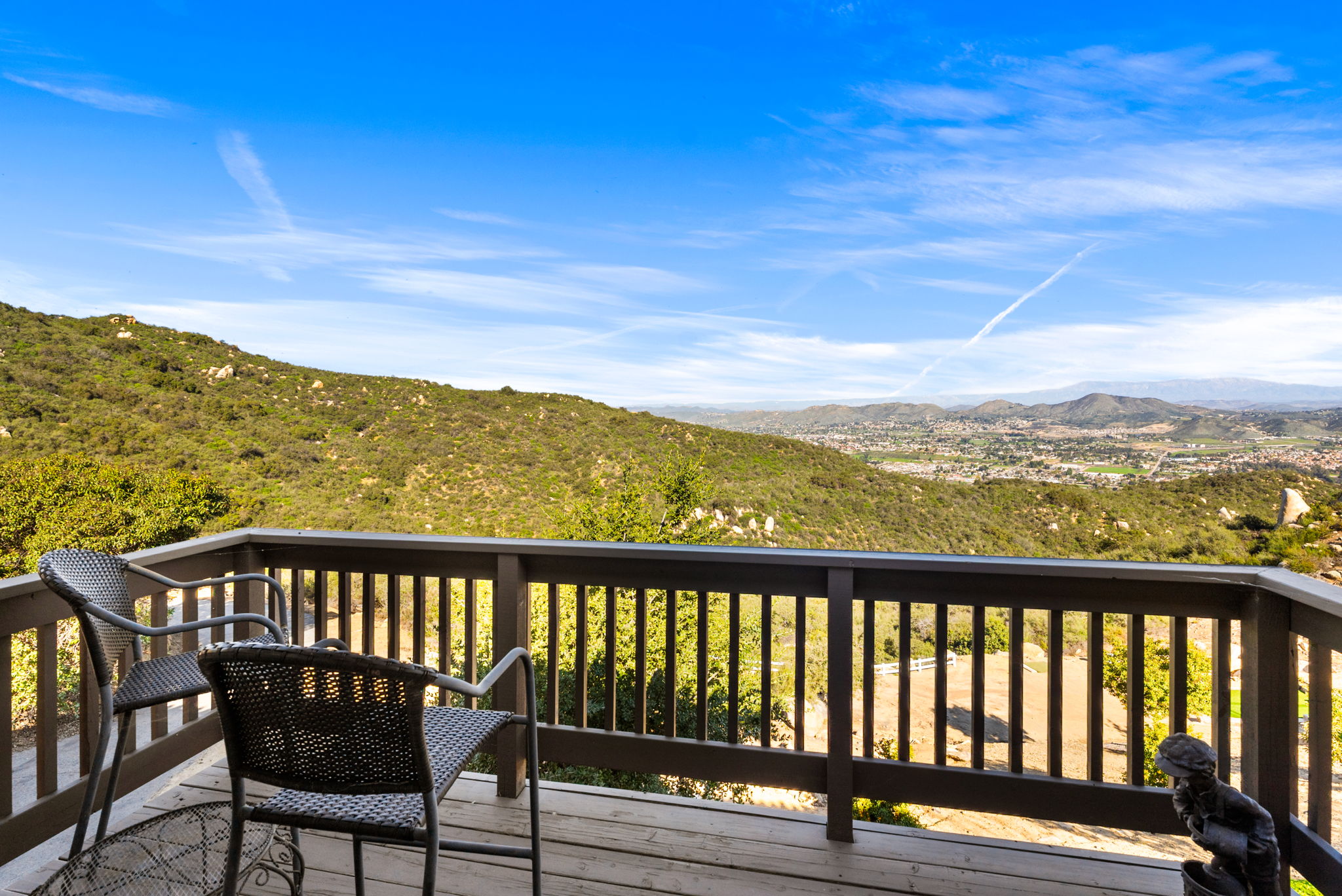 This spacious wooden deck offers a spectacular panoramic view of the rolling hills and the distant town below under a clear blue sky. The area is furnished with comfortable outdoor seating, making it an ideal spot for relaxation and entertaining. The deck features a sturdy wooden railing and overlooks lush landscape, emphasizing a peaceful and scenic outdoor living experience.