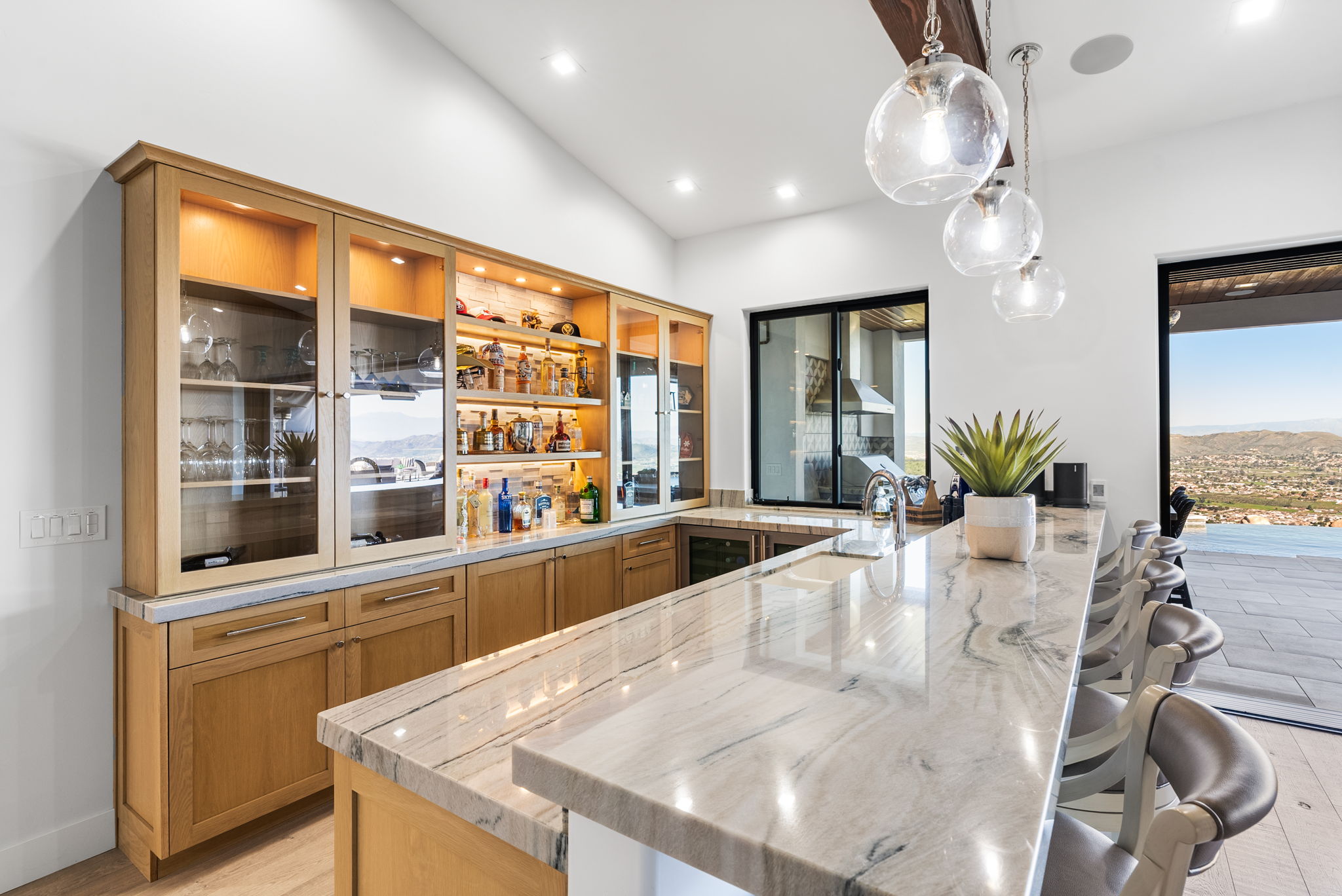 This sophisticated home bar features elegant light oak cabinetry, polished marble countertops, and illuminated open shelving for display. The space is accented by stylish glass pendant lighting and a seamless transition to an outdoor deck with expansive panoramic views. It is a high-end, functional area designed for entertaining with a built-in sink and wine refrigeration.