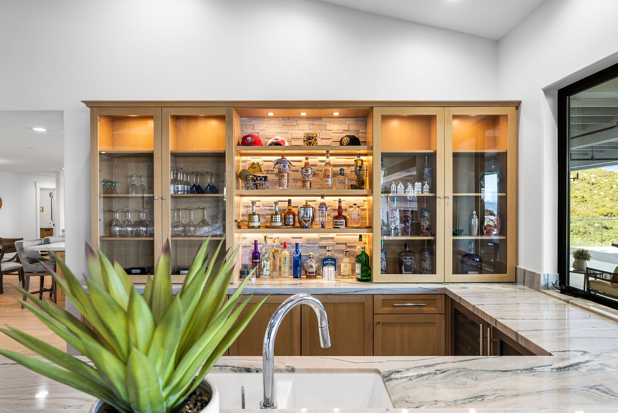 This interior shot showcases a sophisticated home bar area integrated into a kitchen space, featuring custom light wood cabinetry and a marble-patterned countertop. The bar is backlit, displaying an extensive collection of spirits and glassware, creating an elegant focal point. A large, realistic agave plant in the foreground adds a touch of natural color, while a nearby large window reveals a scenic, lush outdoor view.