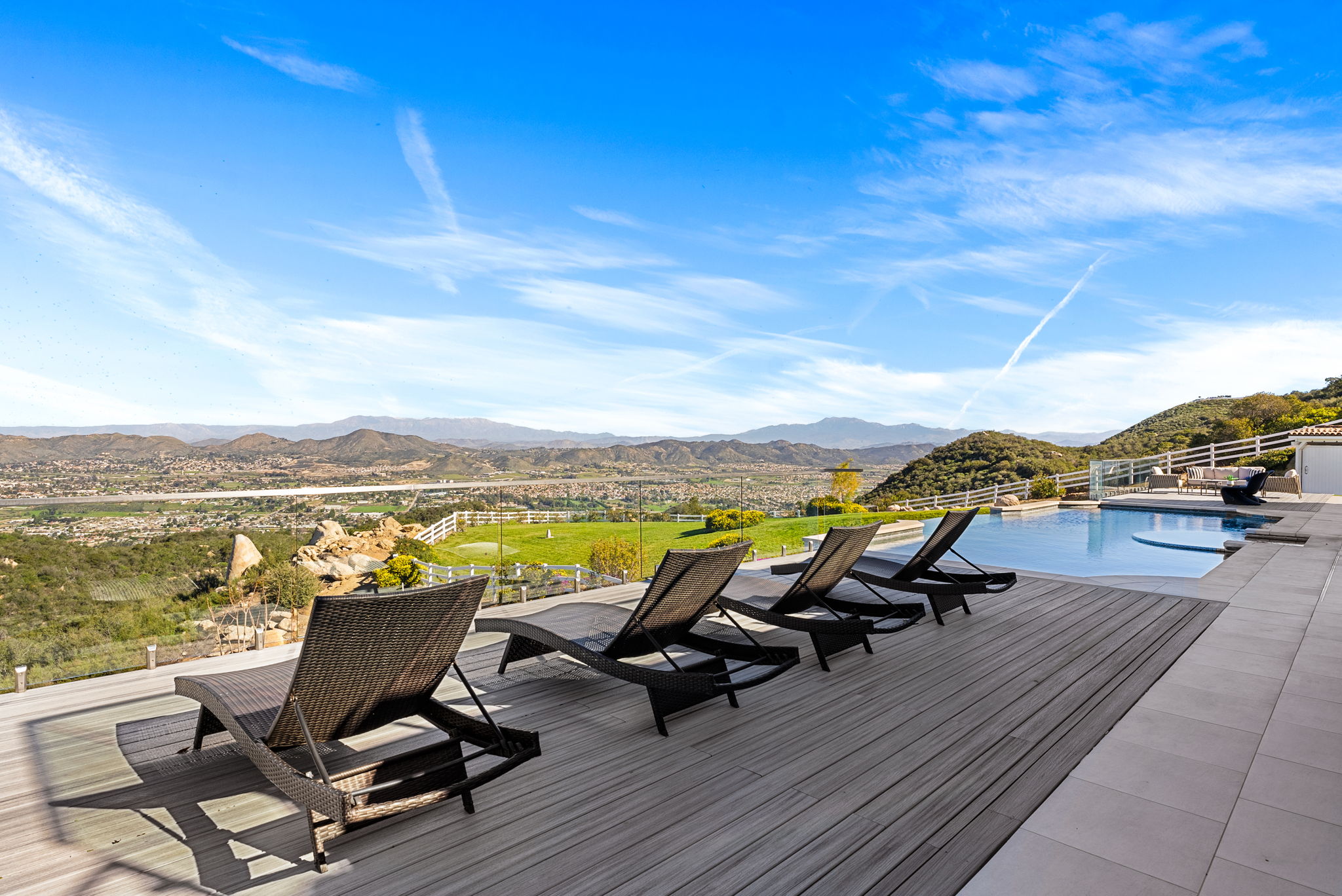This stunning outdoor deck features four contemporary wicker lounge chairs arranged perfectly for soaking in the breathtaking panoramic mountain and valley views. The space seamlessly integrates a sleek infinity-edge swimming pool with an elegant glass railing, creating a sophisticated and luxurious resort-style atmosphere ideal for relaxation and entertaining.