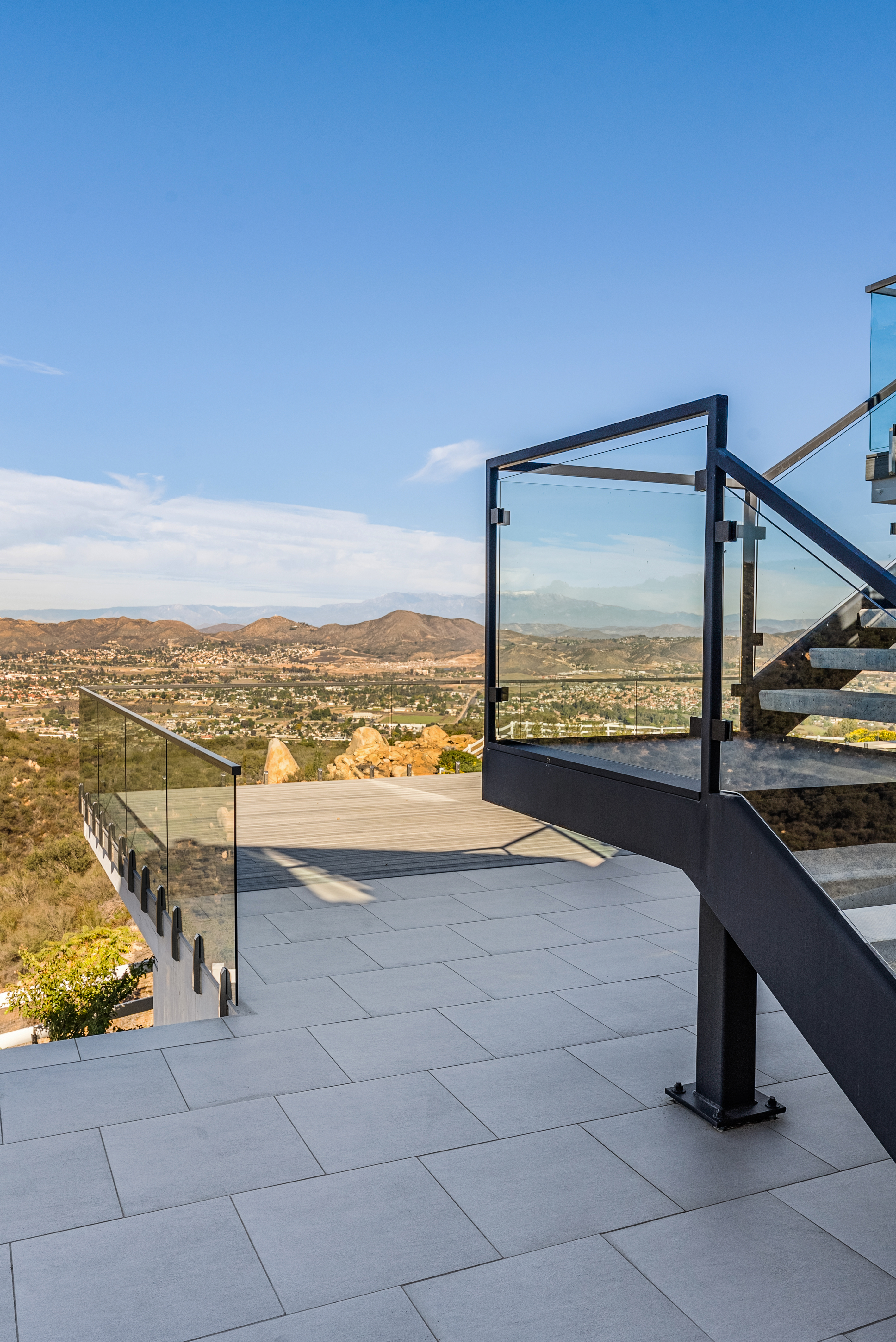 This stunning outdoor balcony features sleek, modern glass railings with black framing, offering an unobstructed, panoramic view of the valley below and surrounding hills. The space is paved with large, light-colored tiles and includes a section of wooden decking, creating a sophisticated and airy aesthetic perfect for relaxation. The design emphasizes seamless integration with the natural landscape, highlighting the property's elevated and scenic location.