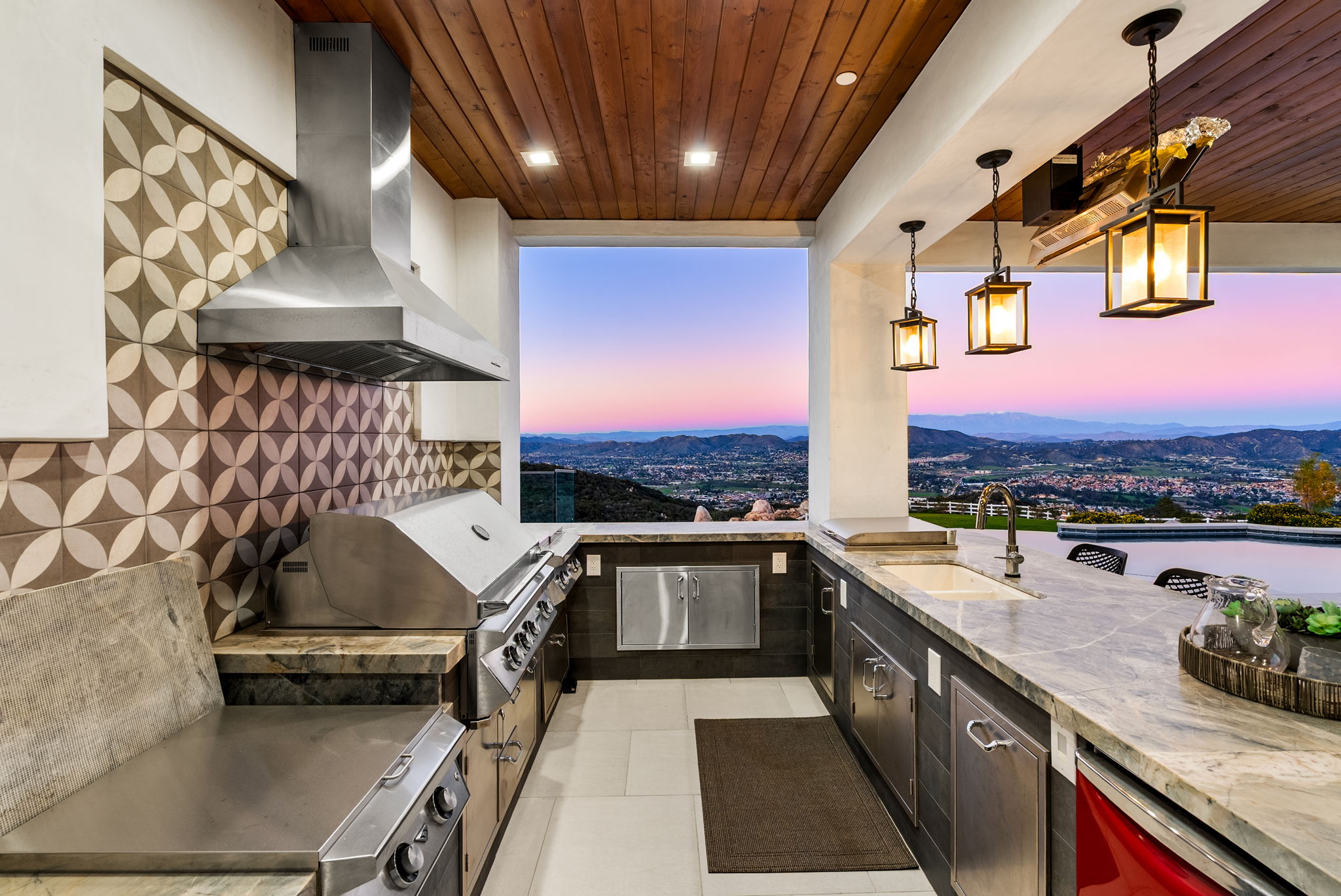 This luxurious outdoor kitchen features high-end stainless steel appliances, stone countertops, and stylish patterned backsplash tiles, all sheltered under a warm wood-plank ceiling. Pendant lighting hangs over a functional counter, while the open-air design frames a stunning, panoramic view of the valley and mountains at sunset. It is an ideal space for sophisticated outdoor entertaining and alfresco dining.