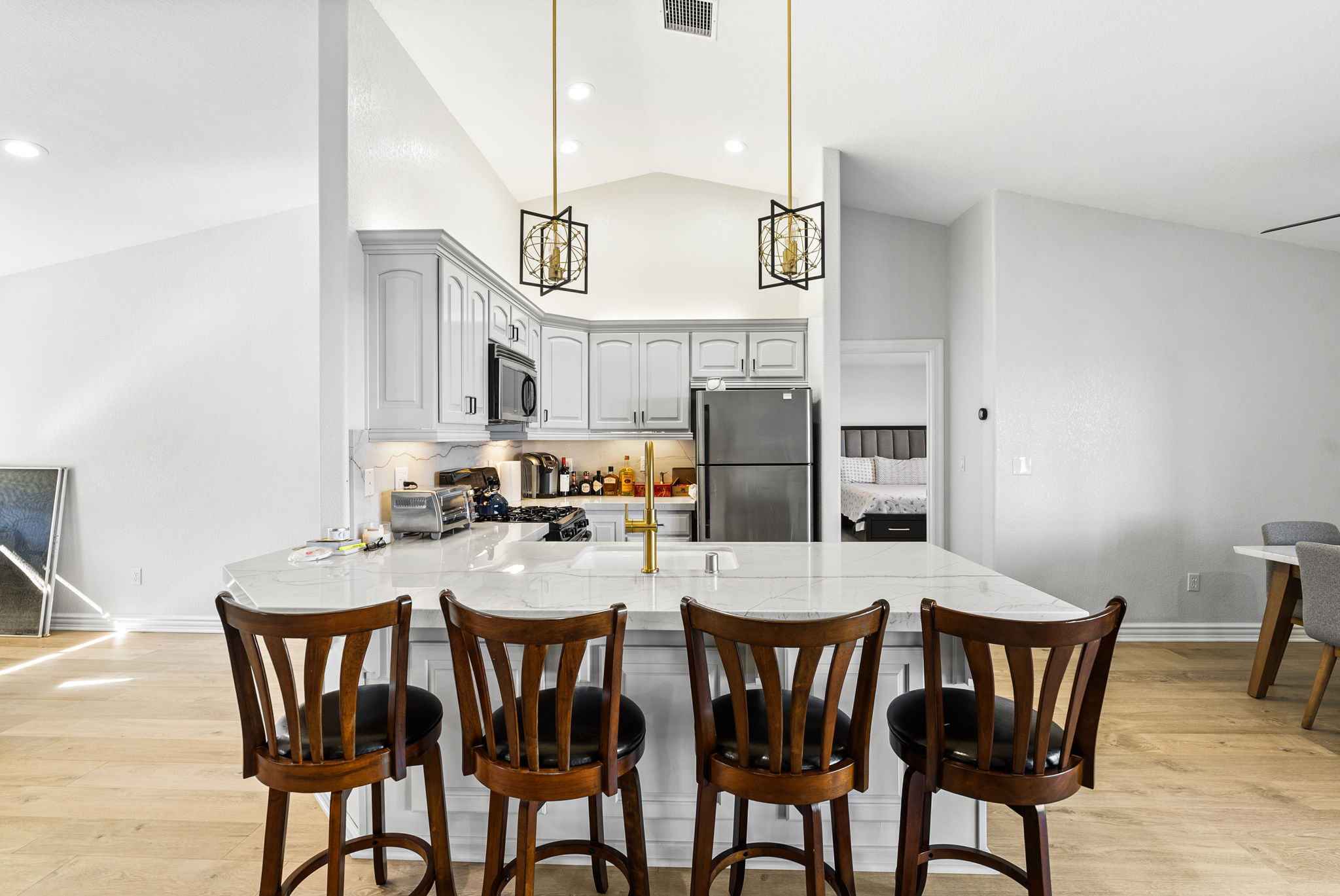 This kitchen features a bright and open design with white cabinetry, a large marble-topped island, and modern gold-toned fixtures. It is equipped with stainless steel appliances and illuminated by stylish geometric pendant lights, creating a clean and inviting atmosphere. The space flows seamlessly into the surrounding living areas, making it ideal for both cooking and casual dining.