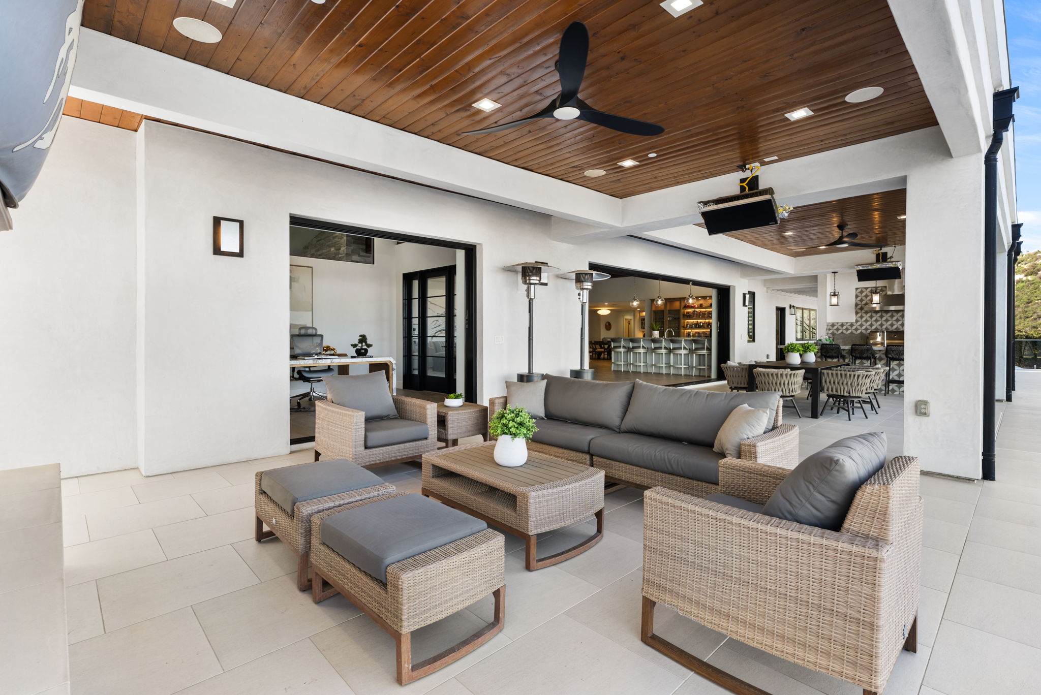 This spacious, luxury outdoor covered patio features a contemporary aesthetic with light stone flooring and a warm wood-plank ceiling. It is furnished with comfortable wicker outdoor seating, a central coffee table, and patio heaters, offering a seamless transition to the interior living spaces. The design emphasizes indoor-outdoor living, perfect for entertainment and relaxation.
