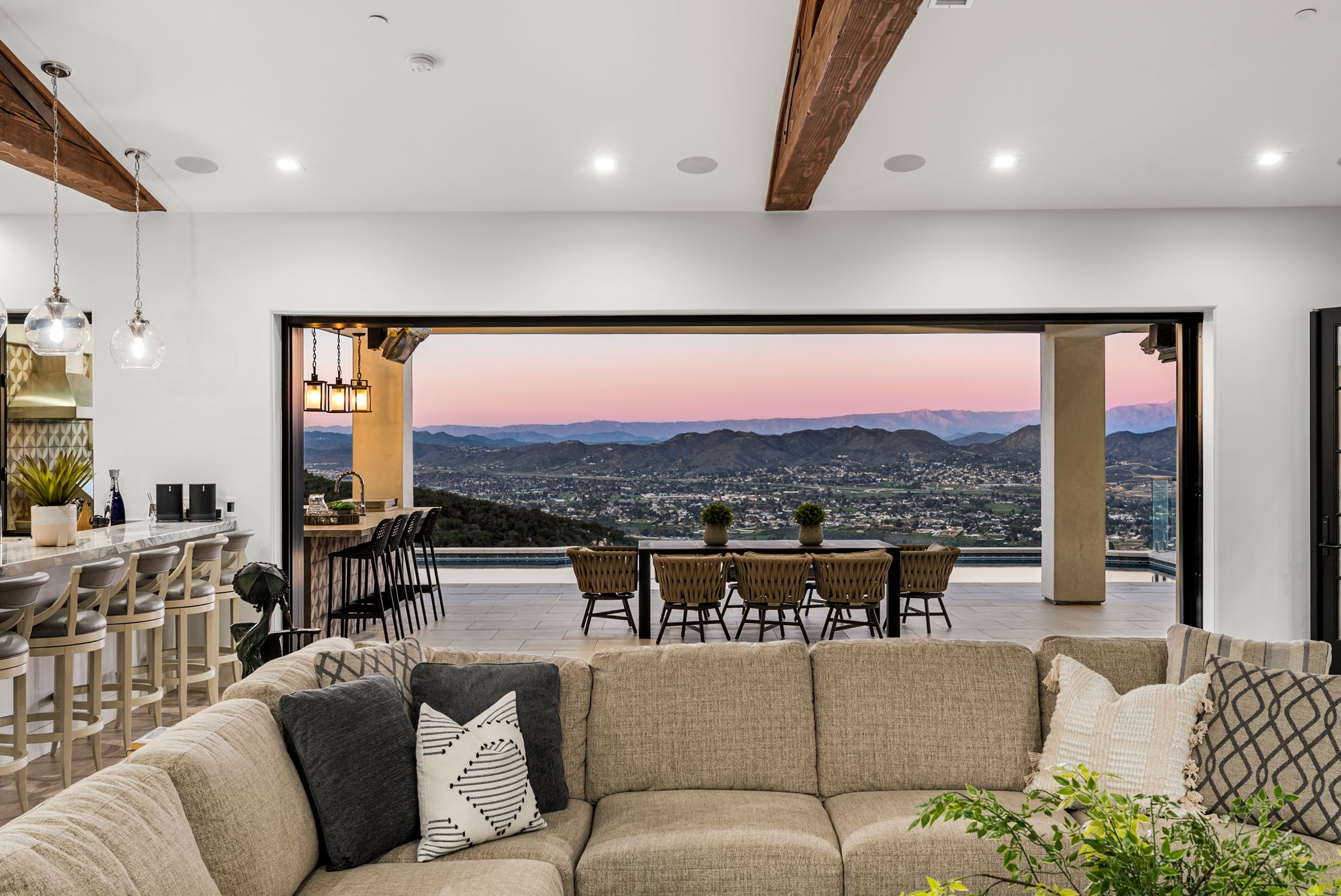 This spacious interior living area features an open floor plan that seamlessly connects to a covered outdoor patio through expansive sliding glass doors. The space highlights a modern, comfortable living room with a neutral-toned sofa, overlooking a stunning sunset view of a valley and mountain range. The design emphasizes indoor-outdoor living, with contemporary furnishings, warm ambient lighting, and high-end materials that create a luxurious and inviting atmosphere.