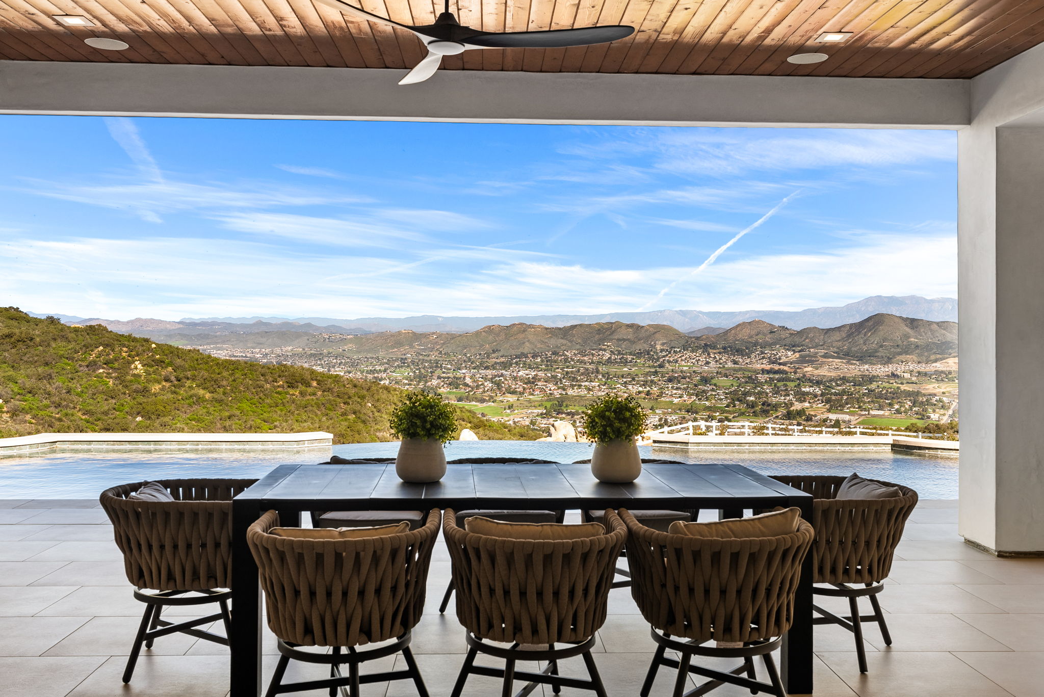 This covered outdoor patio area features a sleek dining set with woven chairs and a central table, perfectly positioned for panoramic mountain and valley views. The space seamlessly transitions into an infinity-edge pool, creating an ideal setting for luxury outdoor entertaining. The modern architecture is enhanced by natural wood ceiling accents and integrated lighting.