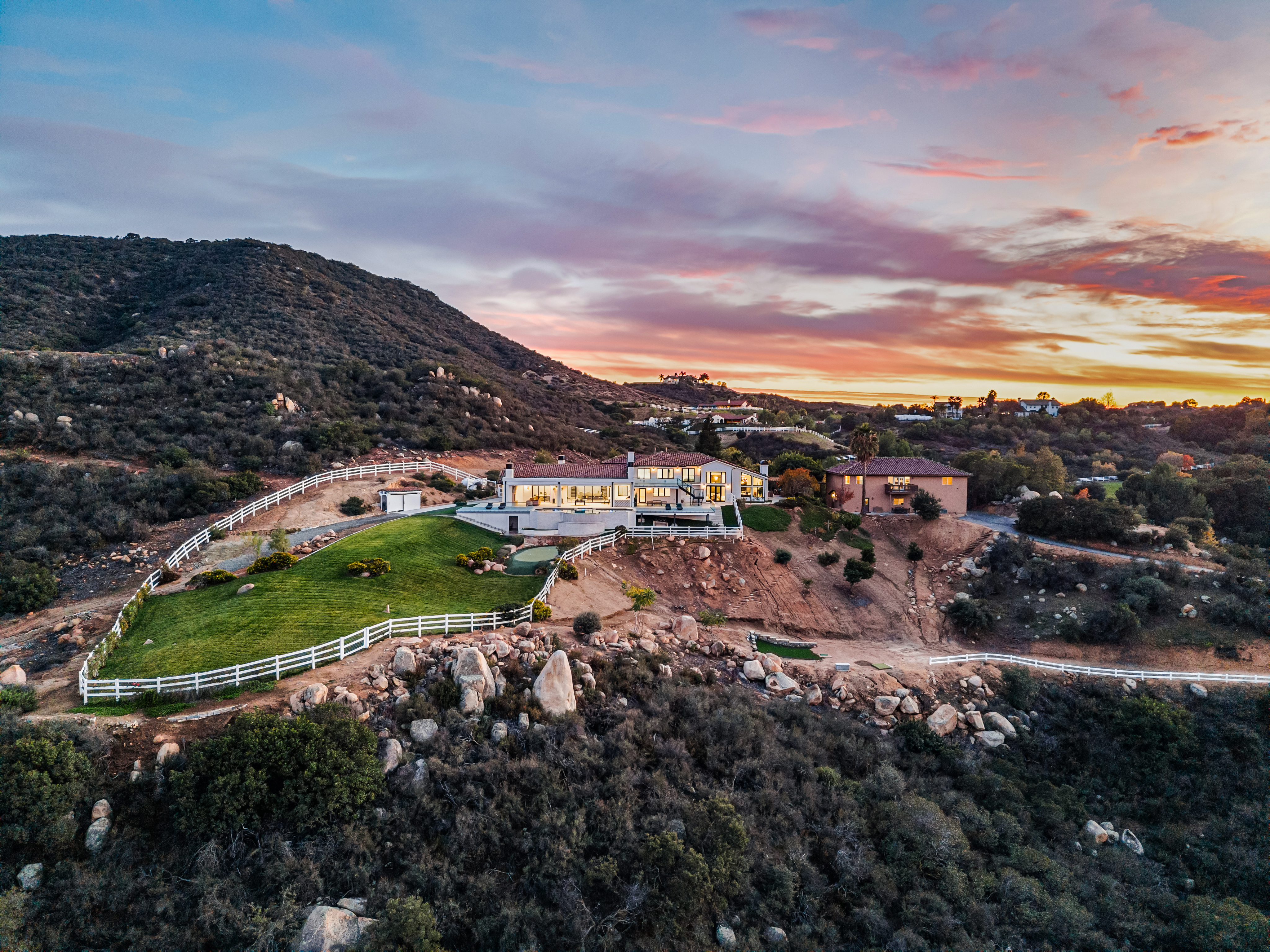 This stunning aerial shot captures a sprawling hilltop estate set against a dramatic, colorful sunset. The property features a large, contemporary home with expansive glass windows, a beautifully manicured green lawn enclosed by white fencing, and rugged, naturalistic landscaping that blends seamlessly into the surrounding terrain. The elevated vantage point highlights the home's privacy, architectural appeal, and the impressive scenic landscape of the surrounding hills.