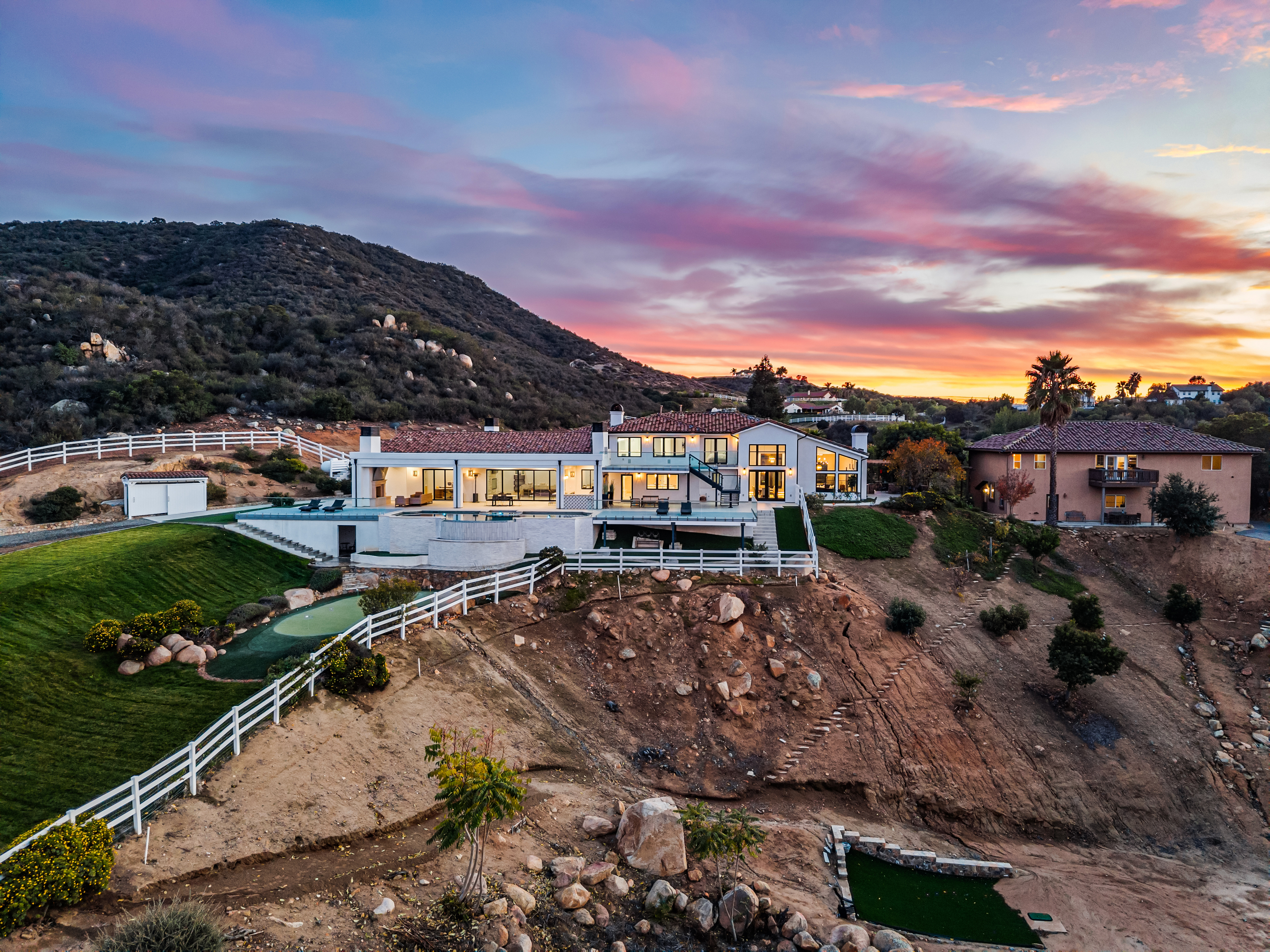 This stunning aerial view captures a luxurious, modern estate perched on a hillside during a vibrant sunset. The property features a pristine pool and spa deck, a manicured lawn with a putting green, and expansive glass doors that emphasize an indoor-outdoor lifestyle. The combination of its elevated position, contemporary architecture, and scenic backdrop makes this a highly desirable, private residence.