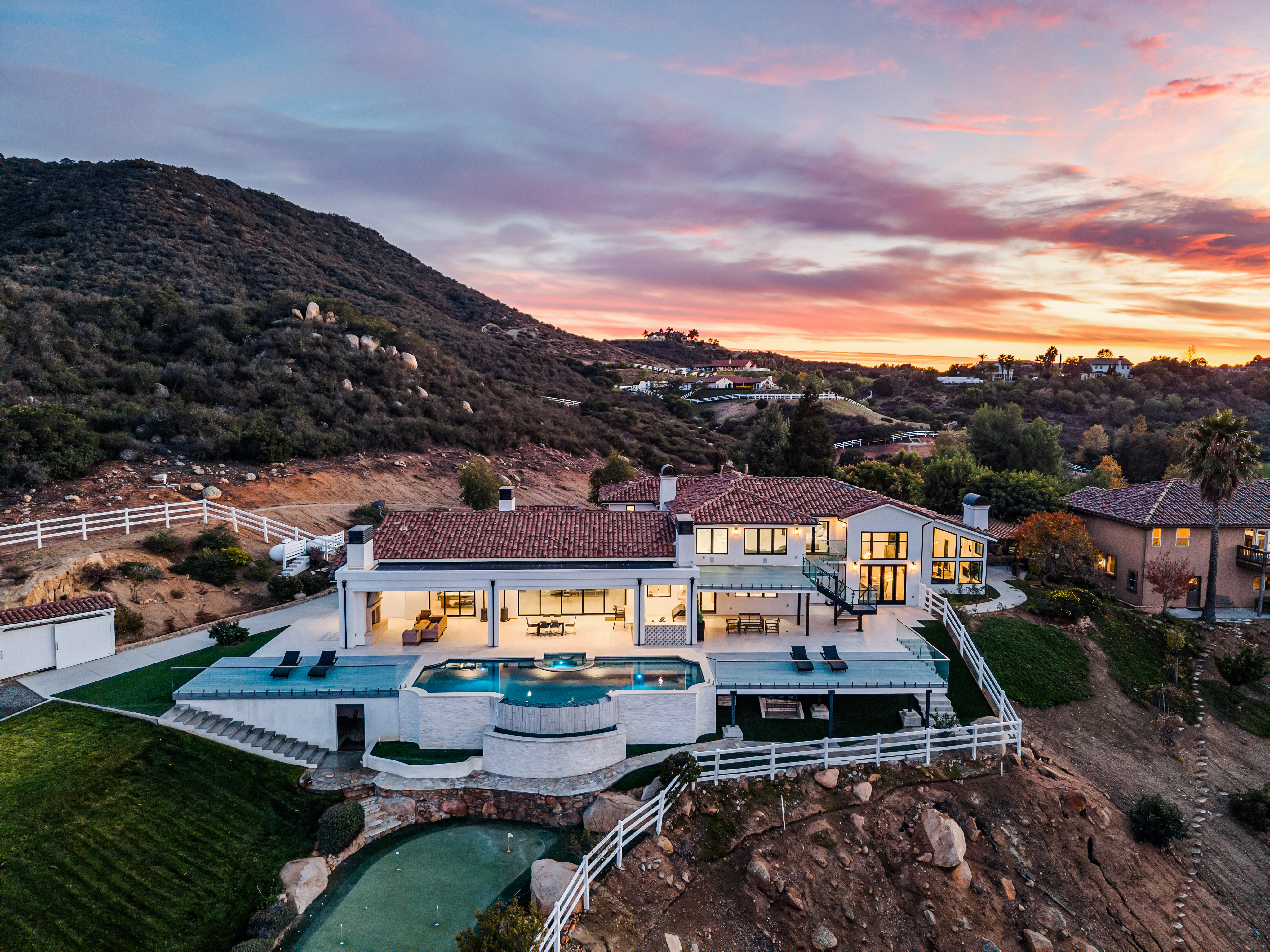 This stunning aerial view captures a contemporary luxury estate nestled against a rugged hillside at sunset. The property features a large infinity-edge pool, multiple spacious terraces, and a private putting green, all seamlessly blending indoor and outdoor living spaces. The warm illumination of the house contrasts beautifully with the colorful sky and natural landscape, emphasizing its premium design and exclusive appeal.