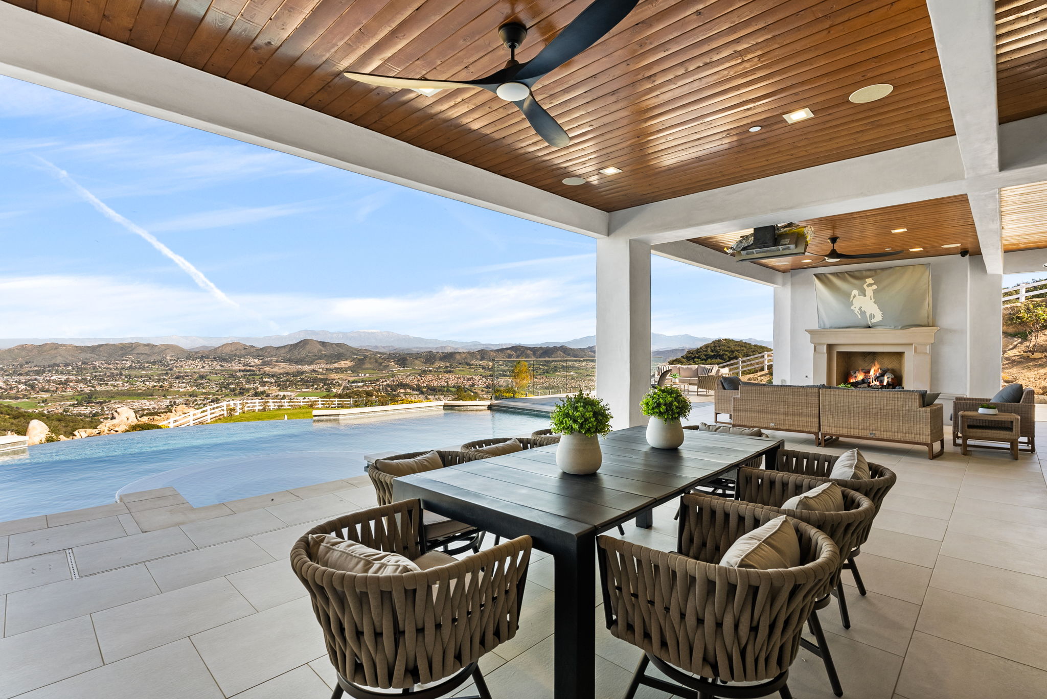 This luxurious covered patio features an expansive infinity pool overlooking a stunning panoramic mountain and valley vista. The space includes a contemporary dining set, comfortable outdoor lounge furniture, and a built-in fireplace, making it an ideal setting for high-end outdoor entertaining. The modern architecture, highlighted by wood-paneled ceilings and clean lines, emphasizes a seamless transition between indoor and outdoor living.