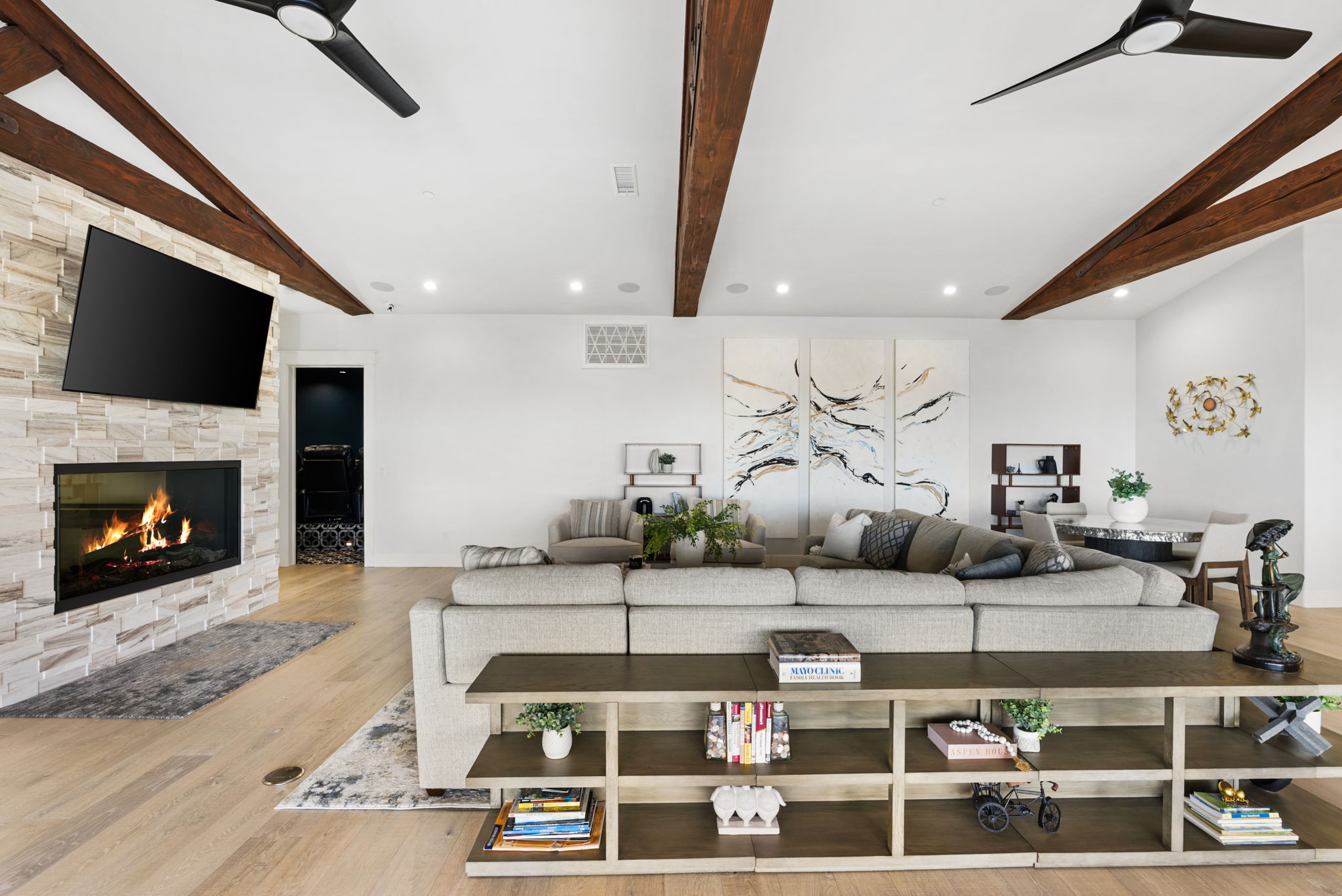 This spacious, open-concept living area features a large sectional sofa facing a stunning stone-clad fireplace and a mounted television. The room is accented by dramatic dark wood exposed ceiling beams, light hardwood flooring, and a decorative console table that provides additional storage and display space. The neutral color palette and artistic wall decor create a modern, inviting, and sophisticated atmosphere.