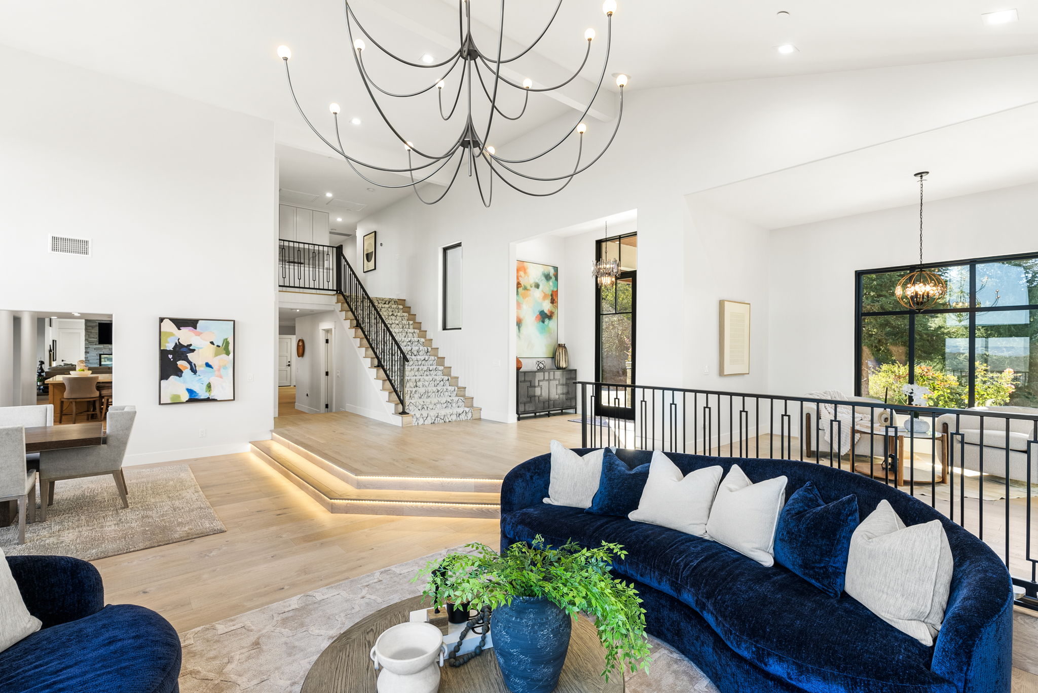 This spacious, modern open-concept living area features a dramatic, multi-armed chandelier and a sophisticated deep blue curved velvet sofa. The room showcases high ceilings, elegant light wood flooring with integrated step lighting, and a grand staircase with a custom patterned runner. Large windows and refined contemporary artwork create a bright, airy, and high-end aesthetic perfect for luxury living.