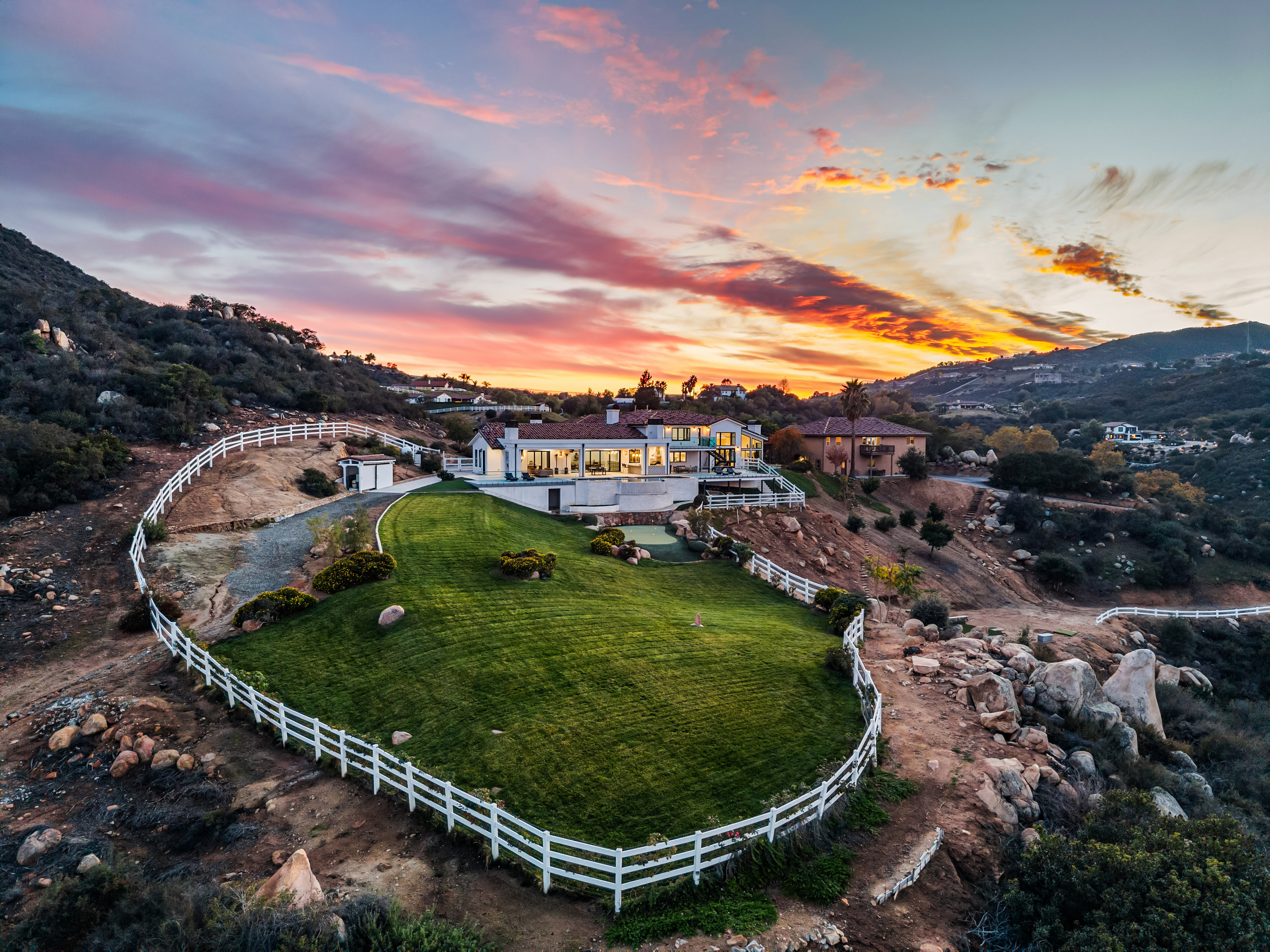 This stunning aerial view captures a luxurious modern estate perched on a hillside at sunset, highlighted by vibrant orange and purple clouds. The property features a large, manicured lawn enclosed by a classic white fence, a spacious back patio with a pool area, and a multi-level architectural design. The surrounding landscape is rugged and natural, creating a beautiful contrast with the polished, well-lit residential home.