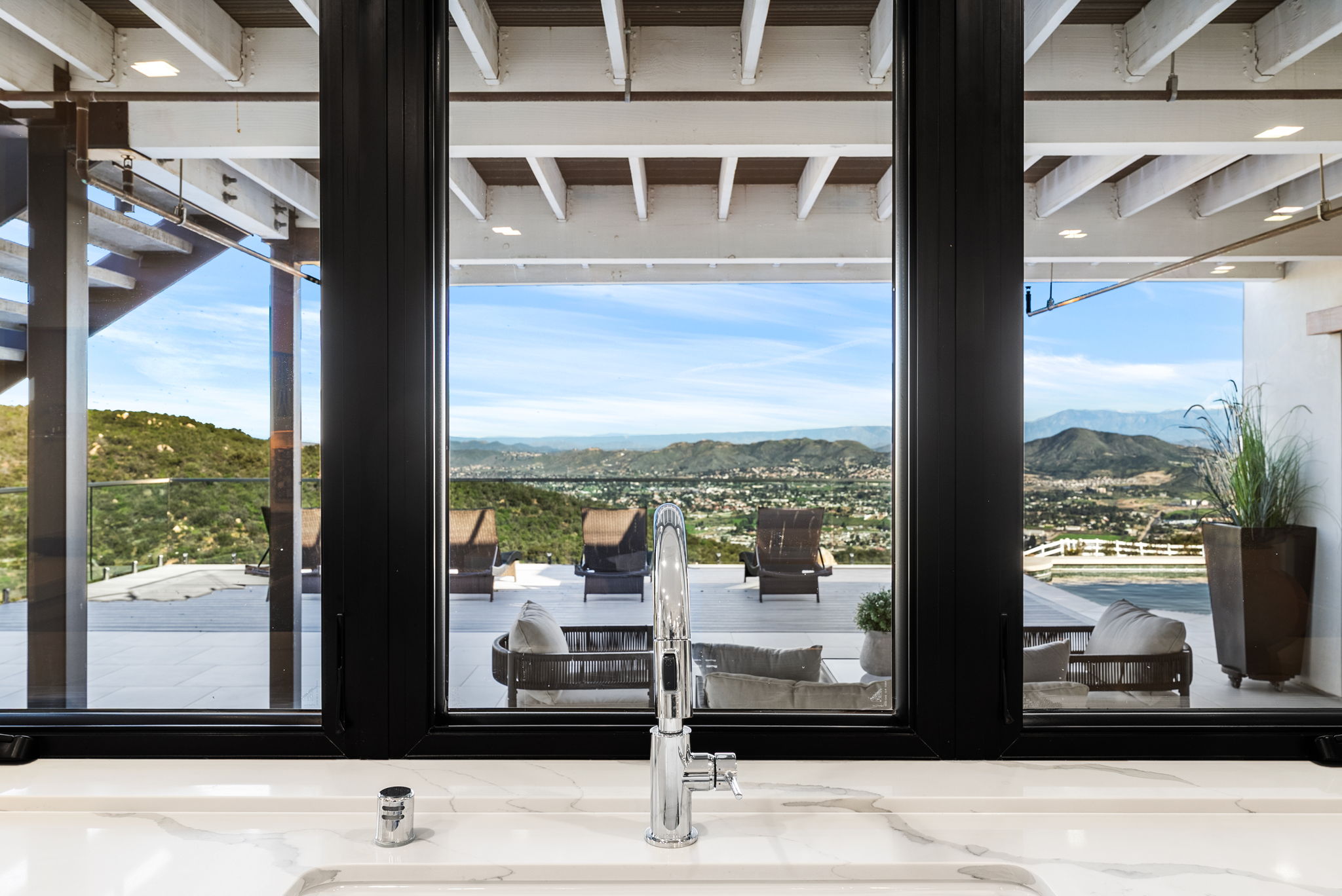 This interior kitchen view features a modern, high-end chrome faucet set against a sleek white marble countertop. The large, black-framed windows provide a striking architectural element that perfectly frames a panoramic view of an expansive outdoor patio and the rolling hills beyond. The overall aesthetic is one of refined luxury and seamless indoor-outdoor living, ideal for a high-end property listing.