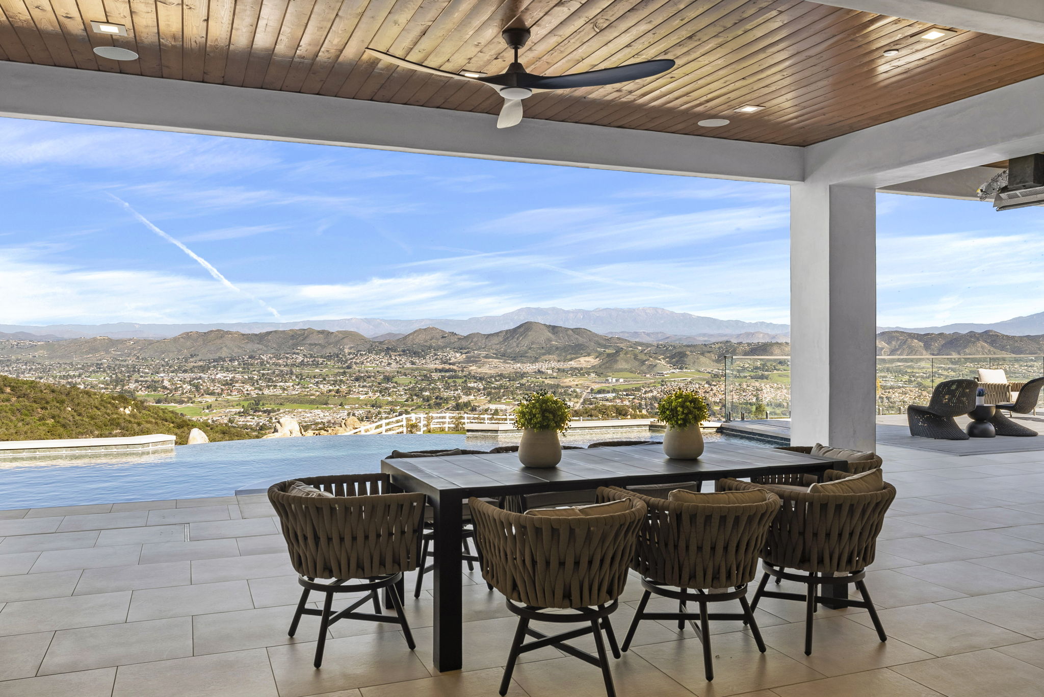 This luxurious covered patio features an elegant outdoor dining set with woven chairs and a sleek table, perfectly positioned to take advantage of the expansive panoramic valley and mountain views. The space seamlessly transitions to an infinity-edge pool, creating a sophisticated and serene atmosphere for outdoor entertaining. With its high-end ceiling fan, warm wood plank ceiling, and refined tile flooring, the area offers a premier indoor-outdoor living experience.