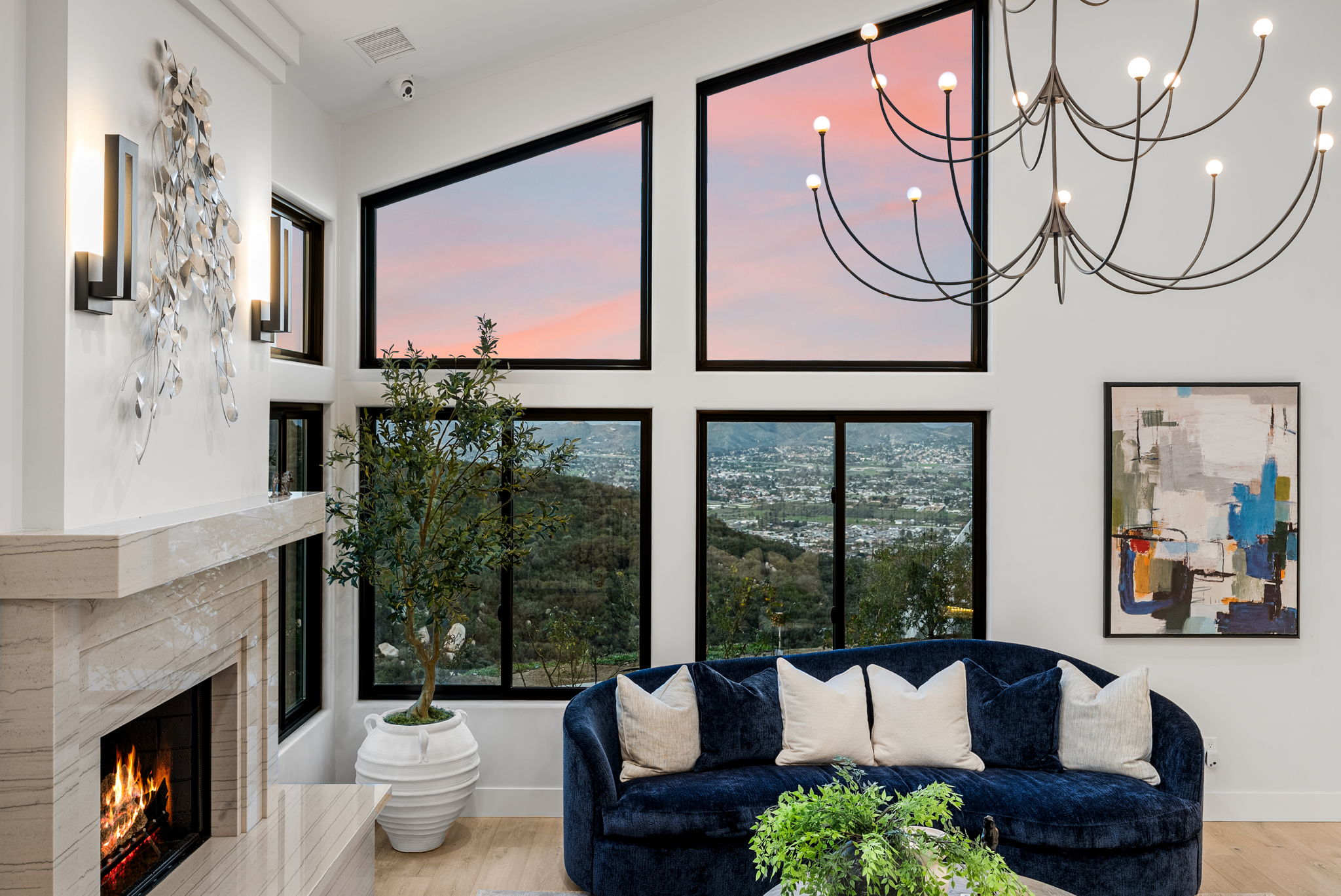 This elegant living room features a plush, deep blue velvet curved sofa and a warm fireplace accented by a large, sculptural metal wall piece. Expansive, custom-shaped windows frame a breathtaking panoramic view of the hillside landscape and valley at sunset. The sophisticated interior is completed by a striking multi-arm chandelier and contemporary abstract artwork, creating an inviting and luxurious atmosphere.