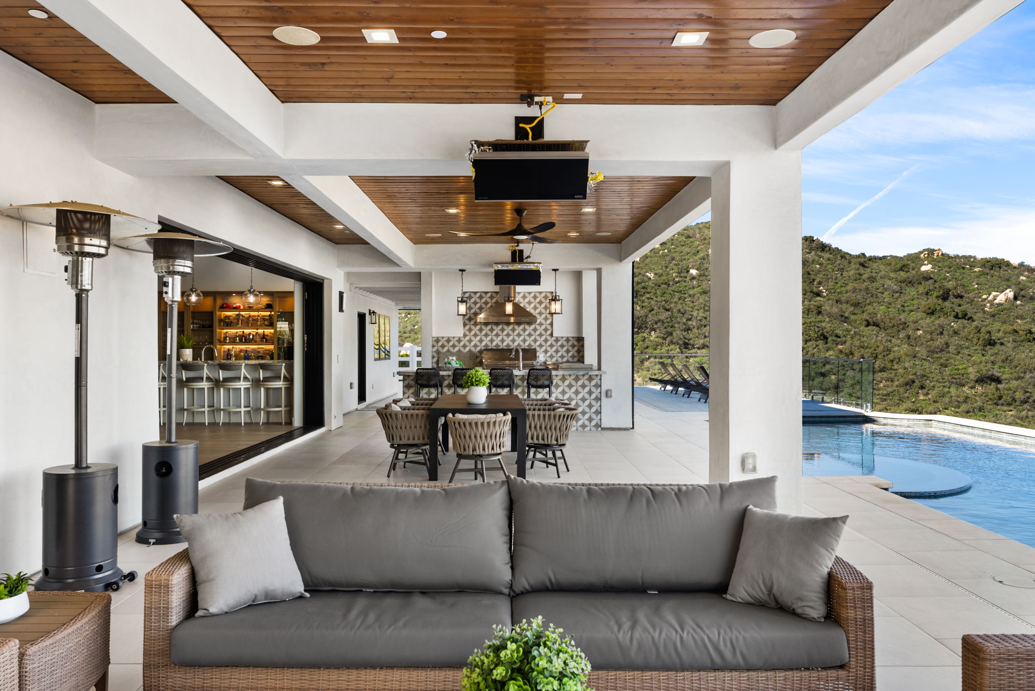 This luxurious covered patio offers a sophisticated outdoor living experience, featuring a sleek, modern dining area and a comfortable lounge space with a grey sofa. The space integrates seamlessly with an outdoor kitchen, complete with a patterned tile backsplash, and opens up to a stunning infinity pool overlooking a scenic, hilly landscape. The high-end design, natural wood ceiling, and seamless transition between indoor and outdoor areas make it an ideal setting for high-end entertaining.