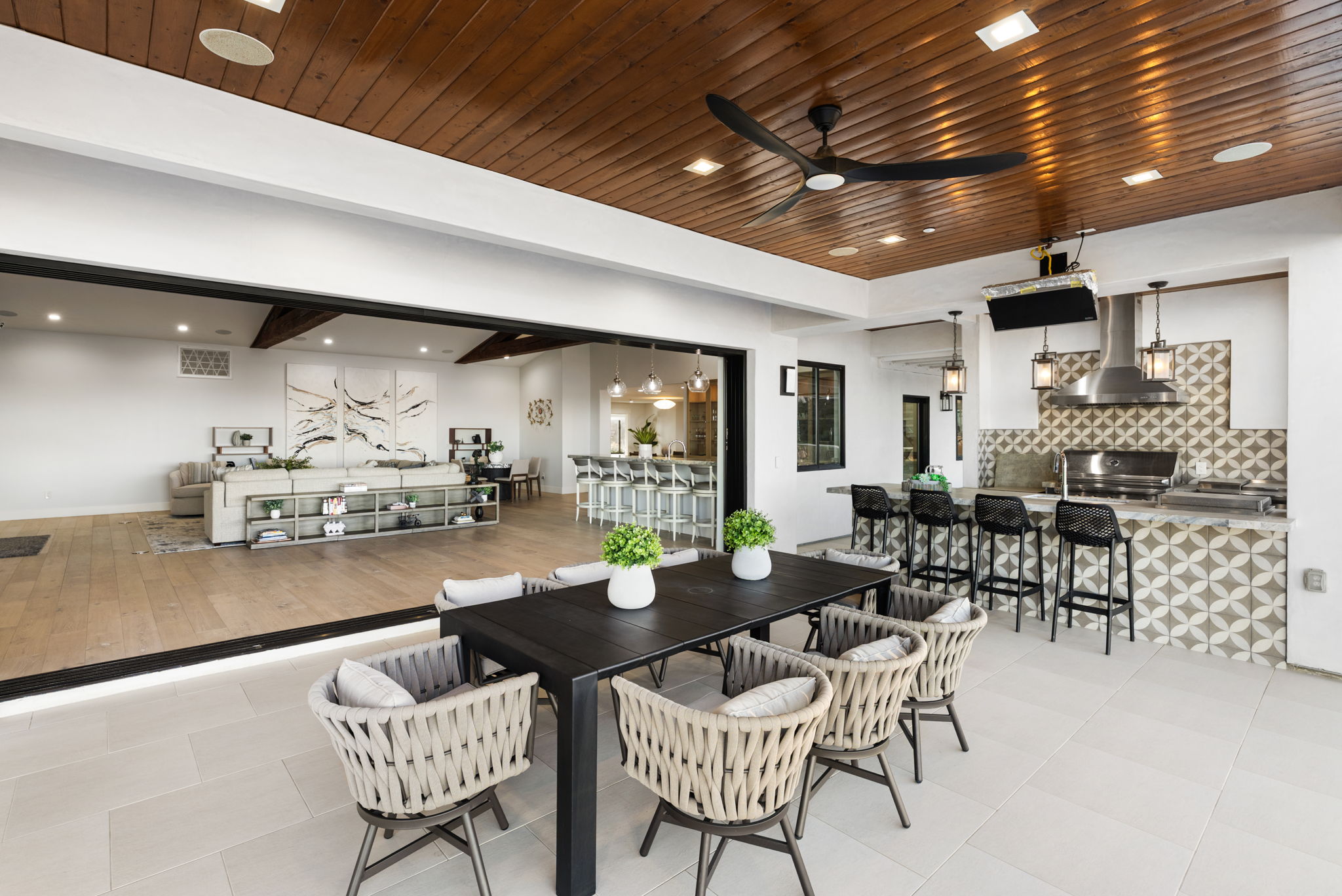 This seamless indoor-outdoor living space features a covered patio equipped with an outdoor kitchen, dining area, and a large ceiling fan. The area transitions effortlessly into an open-concept living and dining room through wide, sliding glass doors, showcasing a sophisticated and airy modern design. High-end finishes, such as patterned tile on the grill station and woven furniture, create a luxurious environment perfect for entertaining.