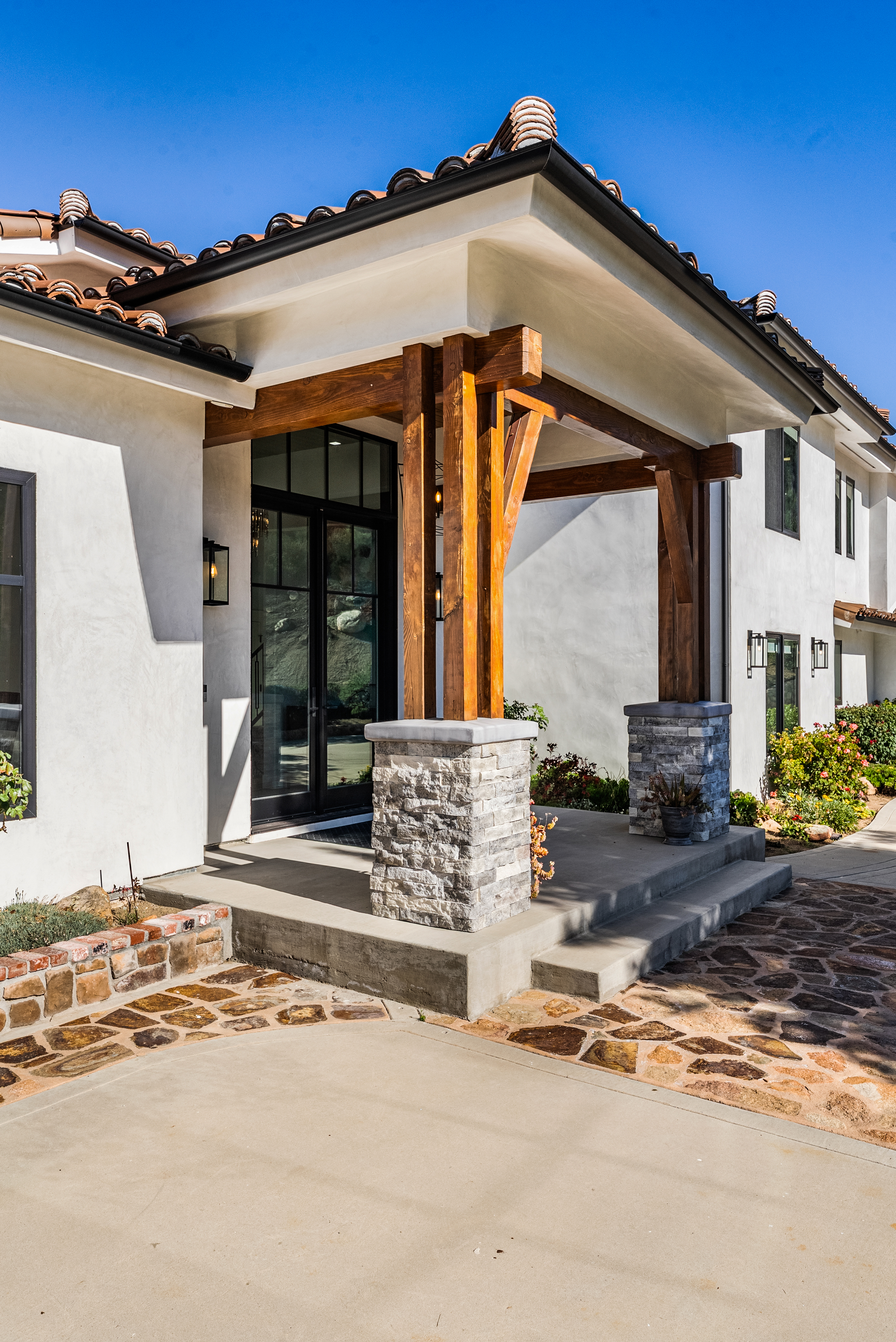 This inviting entryway features a beautiful stucco exterior contrasted with rich, natural wood posts supported by stone-clad bases. The clean lines of the black-framed glass front door and modern sconce lighting enhance the home's contemporary Mediterranean aesthetic. The entrance is further complemented by concrete steps, a stone-accented walkway, and low-maintenance landscaping.