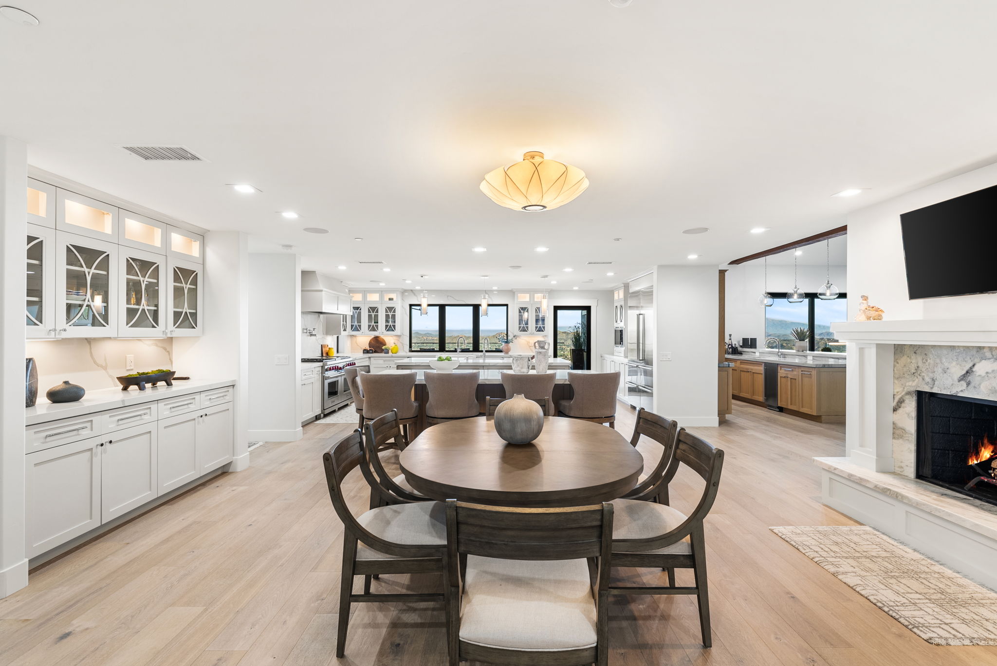 This spacious, open-concept floor plan seamlessly integrates a sophisticated kitchen, dining area, and living space. The kitchen showcases pristine white cabinetry, a large center island with bar seating, and high-end stainless steel appliances, all anchored by light wood flooring. A round dining table serves as a focal point in the foreground, while a stone-faced fireplace adds warmth and elegance to the overall modern aesthetic.