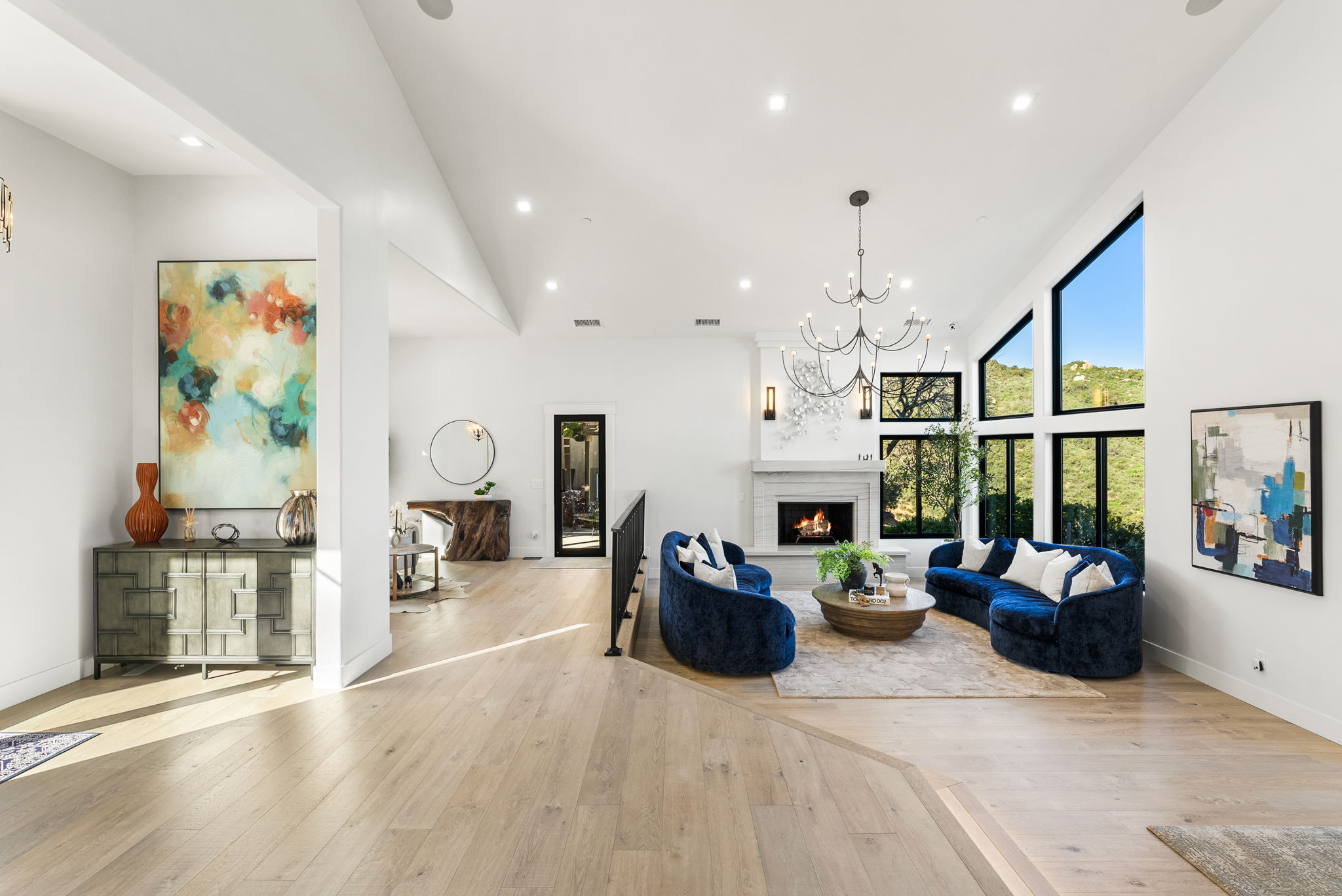 This spacious and light-filled open-concept living room features high vaulted ceilings, elegant hardwood flooring, and striking floor-to-ceiling windows that provide scenic outdoor views. The room is anchored by a cozy fireplace flanked by two deep blue curved sofas, creating a luxurious and inviting atmosphere. Sophisticated artistic accents and a modern chandelier complete the refined, contemporary aesthetic of the home.