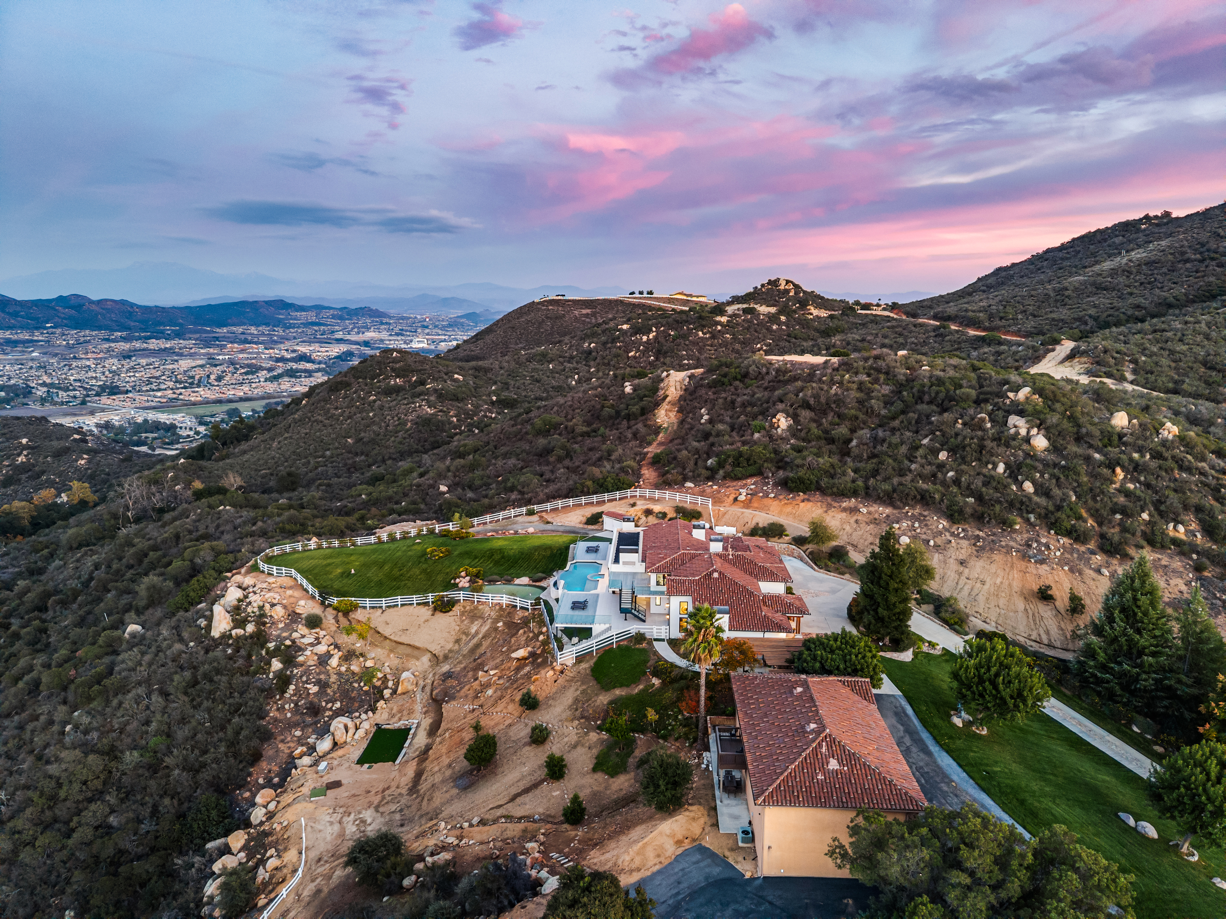 This stunning aerial view captures a sprawling Mediterranean-style estate perched atop a rugged hillside at sunset. The property features a multi-tiered backyard with an inviting swimming pool, manicured lawns, and white fencing, all set against a panoramic backdrop of distant mountains and city lights. The home's classic tile roof and expansive grounds exemplify luxury hilltop living with exceptional views.
