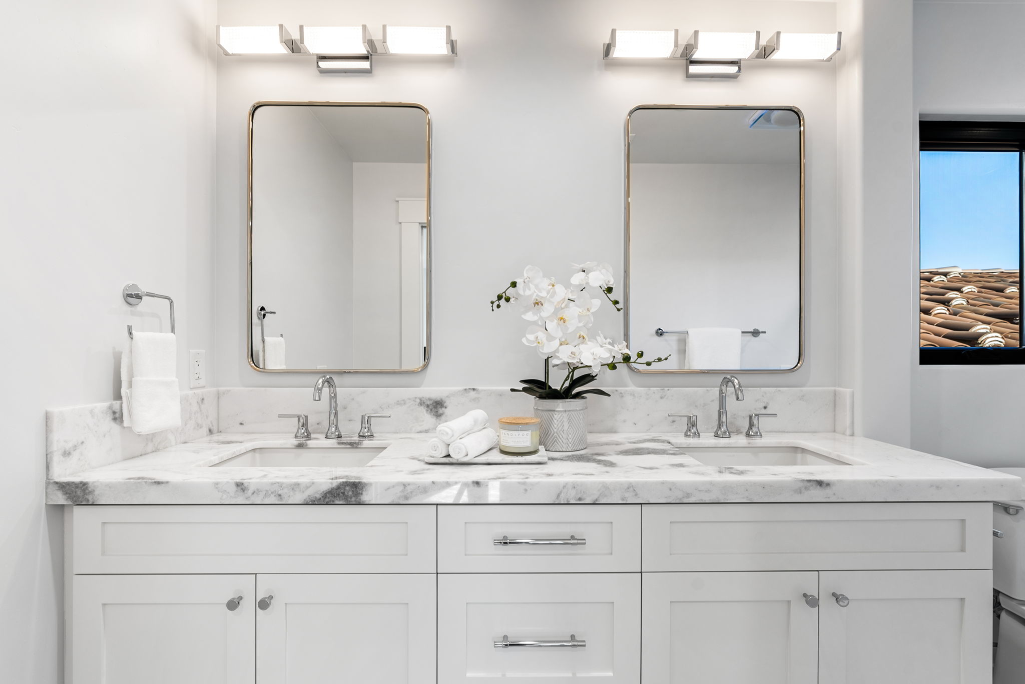This primary bathroom features a clean, modern aesthetic with a double vanity topped by an elegant marble countertop and backsplash. The space is highlighted by two rectangular mirrors with gold frames, matching chrome fixtures, and white shaker-style cabinetry, offering a bright and sophisticated atmosphere.