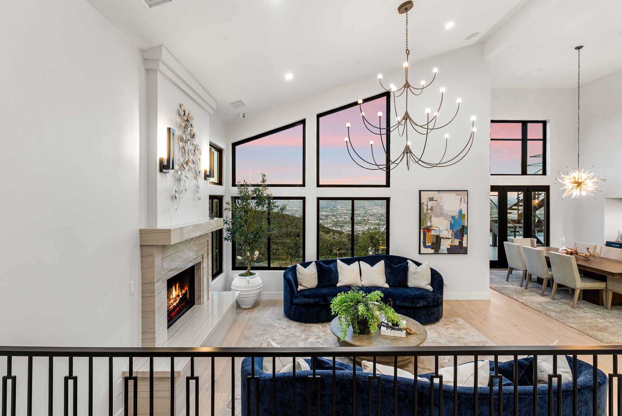 This sophisticated and spacious living room features a dramatic vaulted ceiling with a modern statement chandelier and floor-to-ceiling windows that provide panoramic mountain and valley views. The focal point is a grand stone-clad fireplace, complemented by a luxurious curved deep blue velvet sofa and warm, neutral-toned flooring. The open-concept design seamlessly flows into the adjacent dining area, creating an elegant and airy atmosphere ideal for upscale living.