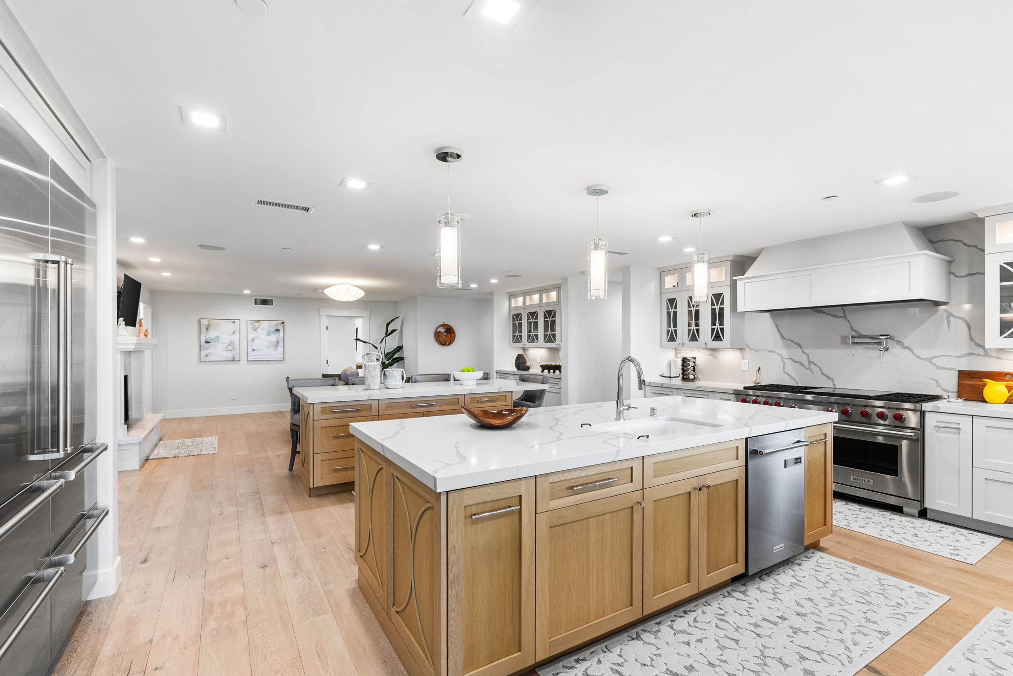 This spacious, modern kitchen features a dual-island layout with warm wood cabinetry and sleek white marble-look quartz countertops. It is equipped with premium stainless steel appliances, including a large Wolf range and a farmhouse-style sink, all illuminated by elegant pendant lighting. The open-concept design seamlessly flows into the adjacent living and dining areas, creating a bright and inviting space perfect for entertaining.