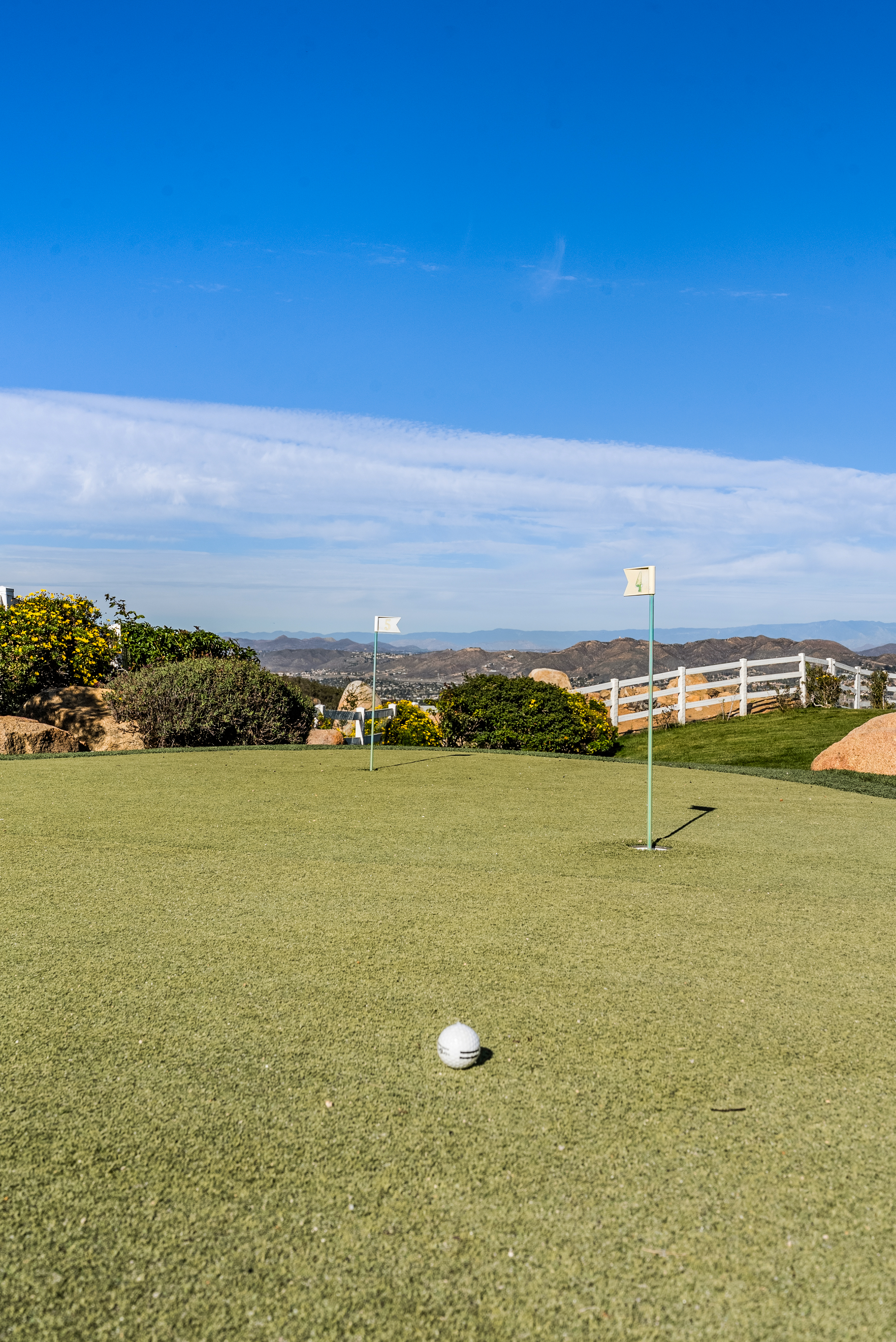 This image showcases a well-maintained putting green amenity featuring artificial turf, multiple holes with flags, and a golf ball positioned in the foreground. Set against a backdrop of scenic rolling hills and a clear blue sky, the area is bordered by native landscaping, large rocks, and a decorative white fence, enhancing its appeal as a premium residential outdoor feature.