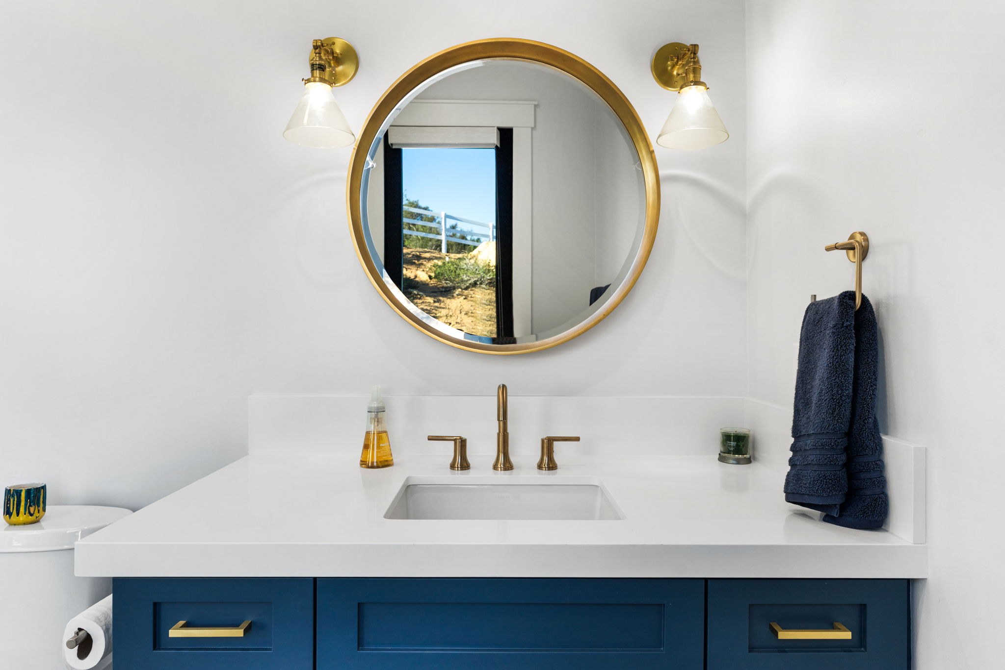 This sleek, modern bathroom features a bold navy blue vanity topped with a crisp white countertop and a contemporary rectangular under-mount sink. Gold-toned fixtures, including the faucet, hardware, and circular mirror frame, create a striking contrast against the clean white walls. A navy blue towel hanging on a matching wall-mounted rack adds a cohesive and stylish touch to the space.