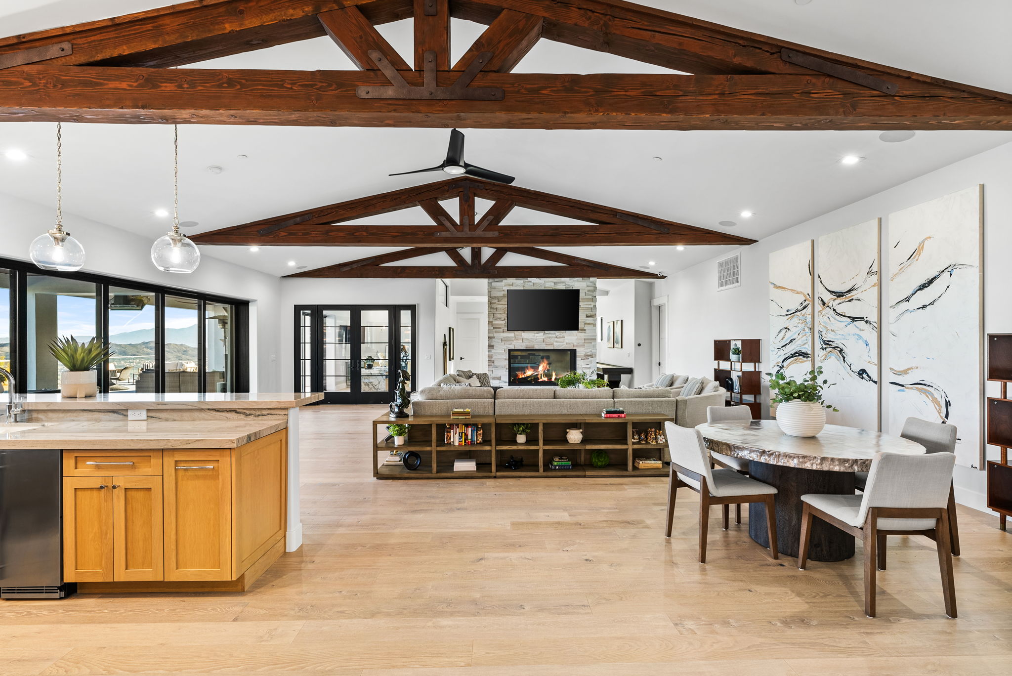 This spacious open-concept living area features stunning exposed wood trusses, light hardwood flooring, and a stone-clad fireplace as a central focal point. The room integrates a dining area, a large sectional sofa, and a kitchen island, creating a cohesive, high-end design suitable for entertaining. Floor-to-ceiling glass doors allow for abundant natural light and seamless indoor-outdoor transition.