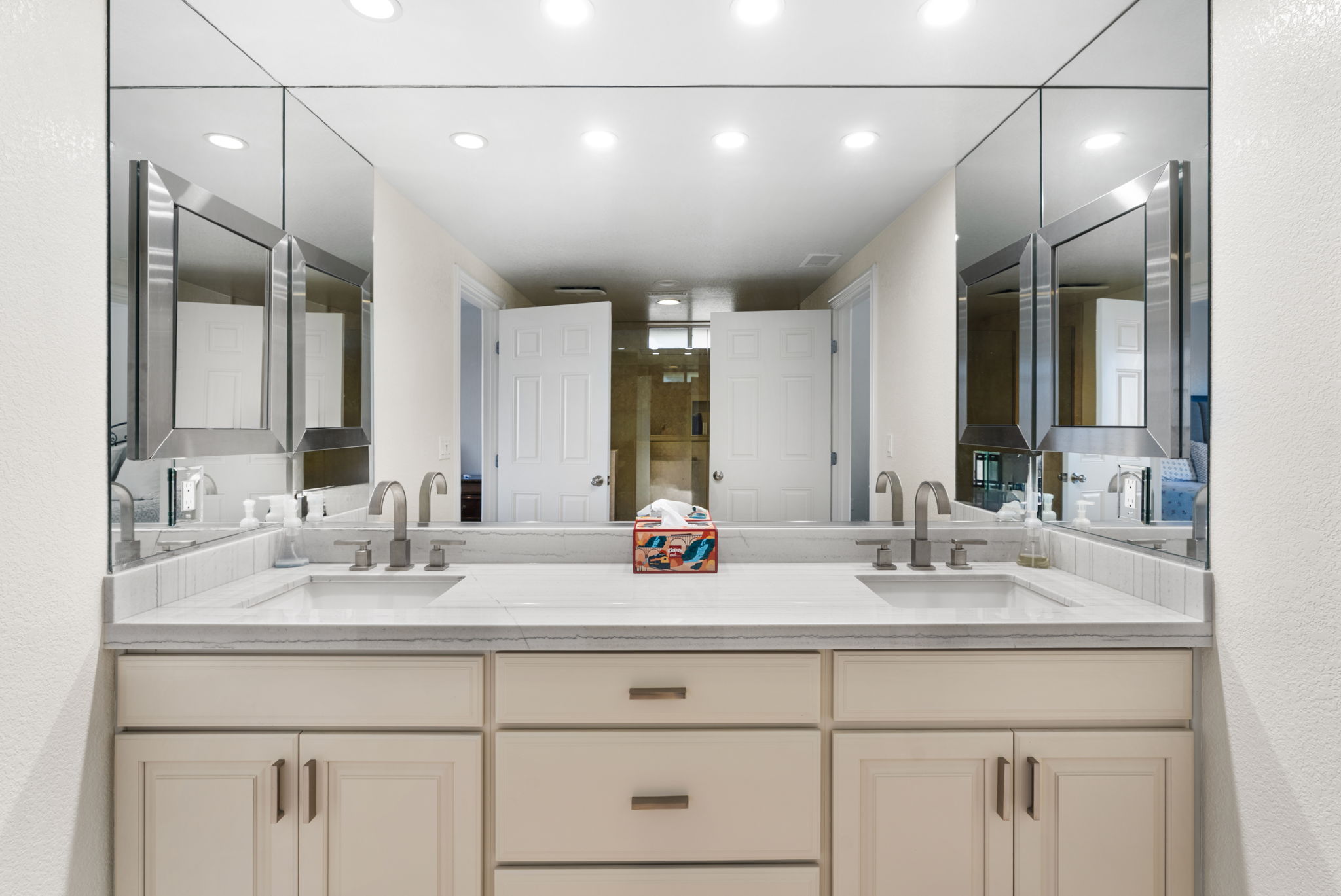 This primary bathroom features a dual-sink vanity with a light-colored stone countertop, modern brushed-nickel faucets, and ample storage cabinetry. A large, expansive mirror spans the wall above the vanity, creating a bright and airy feel, while integrated medicine cabinets offer additional convenience. The clean design and neutral color palette present a functional and well-maintained space.