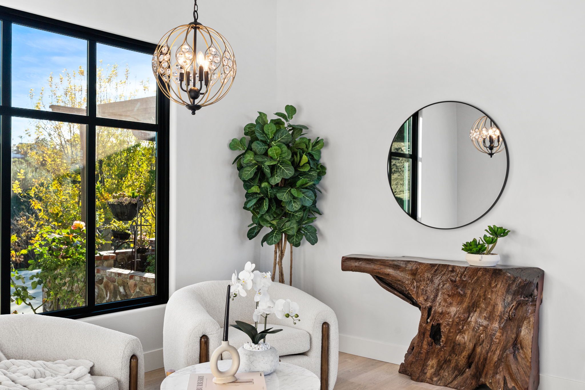 This elegant seating area showcases a contemporary design with two cream-colored bouclé armchairs and a unique, rustic live-edge wood console table. A statement geometric globe chandelier and a large round wall mirror create a sophisticated atmosphere, while a tall artificial tree adds a touch of greenery. The space is brightened by a large floor-to-ceiling window that offers a serene view of the outdoor greenery, blending indoor comfort with nature.