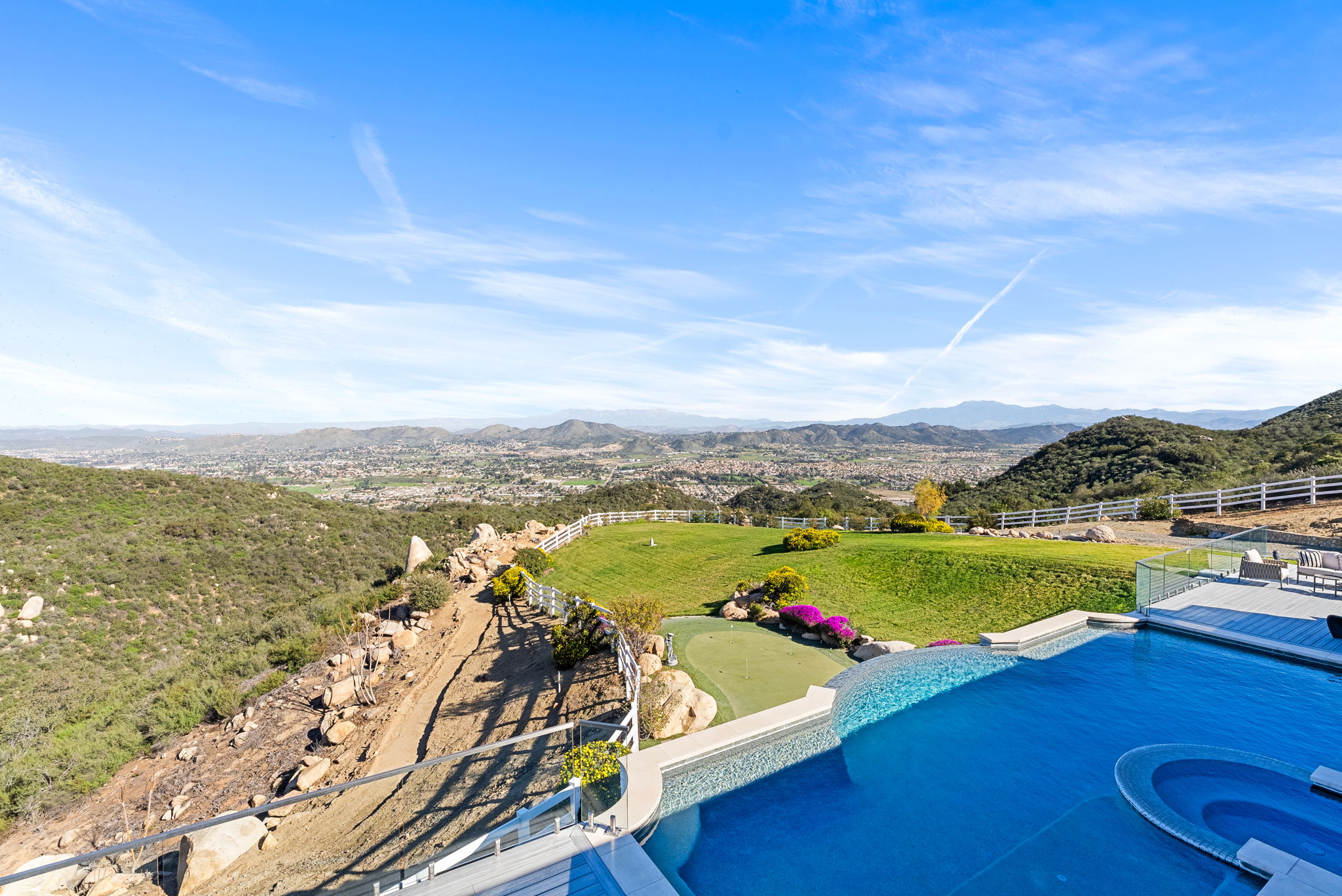 This stunning elevated view captures a luxurious infinity-edge pool and integrated spa overlooking a vast, hilly landscape. The property features a manicured green lawn, a private putting green, and a scenic white fence boundary, all set against a breathtaking mountain backdrop. The combination of high-end outdoor amenities and expansive natural vistas creates a sense of exclusivity and serenity ideal for high-end real estate.