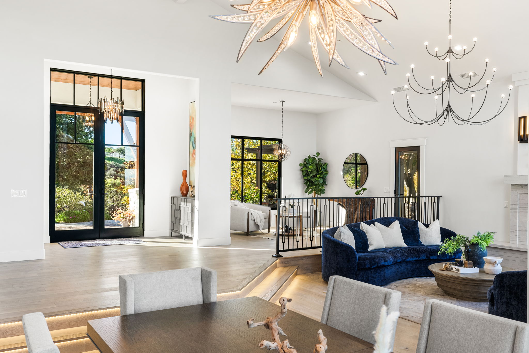 This spacious and elegant open-concept living area features a sophisticated sunken living room with a plush, curved navy velvet sofa. High ceilings with striking, oversized statement chandeliers create a dramatic architectural focal point, while light hardwood floors and large glass-paned doors offer a seamless transition to the outdoors. The space is expertly curated with modern furnishings, balanced by warm natural light, making it ideal for both relaxed living and refined entertaining.