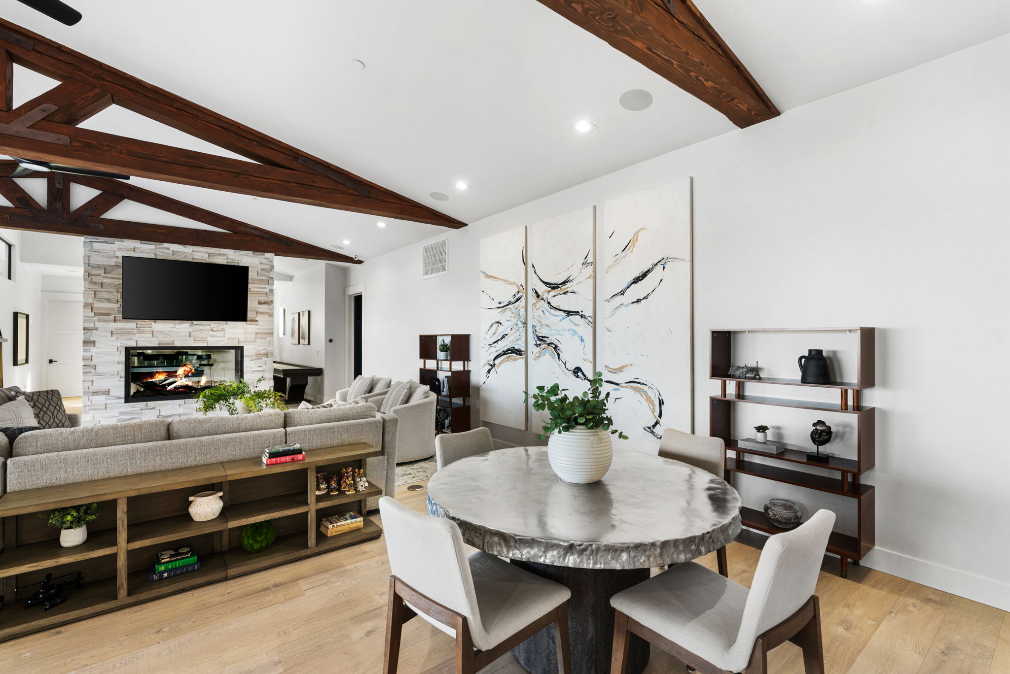This spacious open-concept living area features stunning exposed dark wood beams and a contemporary stone fireplace with an integrated flat-screen television. The space flows seamlessly into a dining area highlighted by a unique textured metallic table and abstract triptych wall art. Light hardwood flooring and neutral-toned furniture create a warm, inviting atmosphere perfect for modern living.