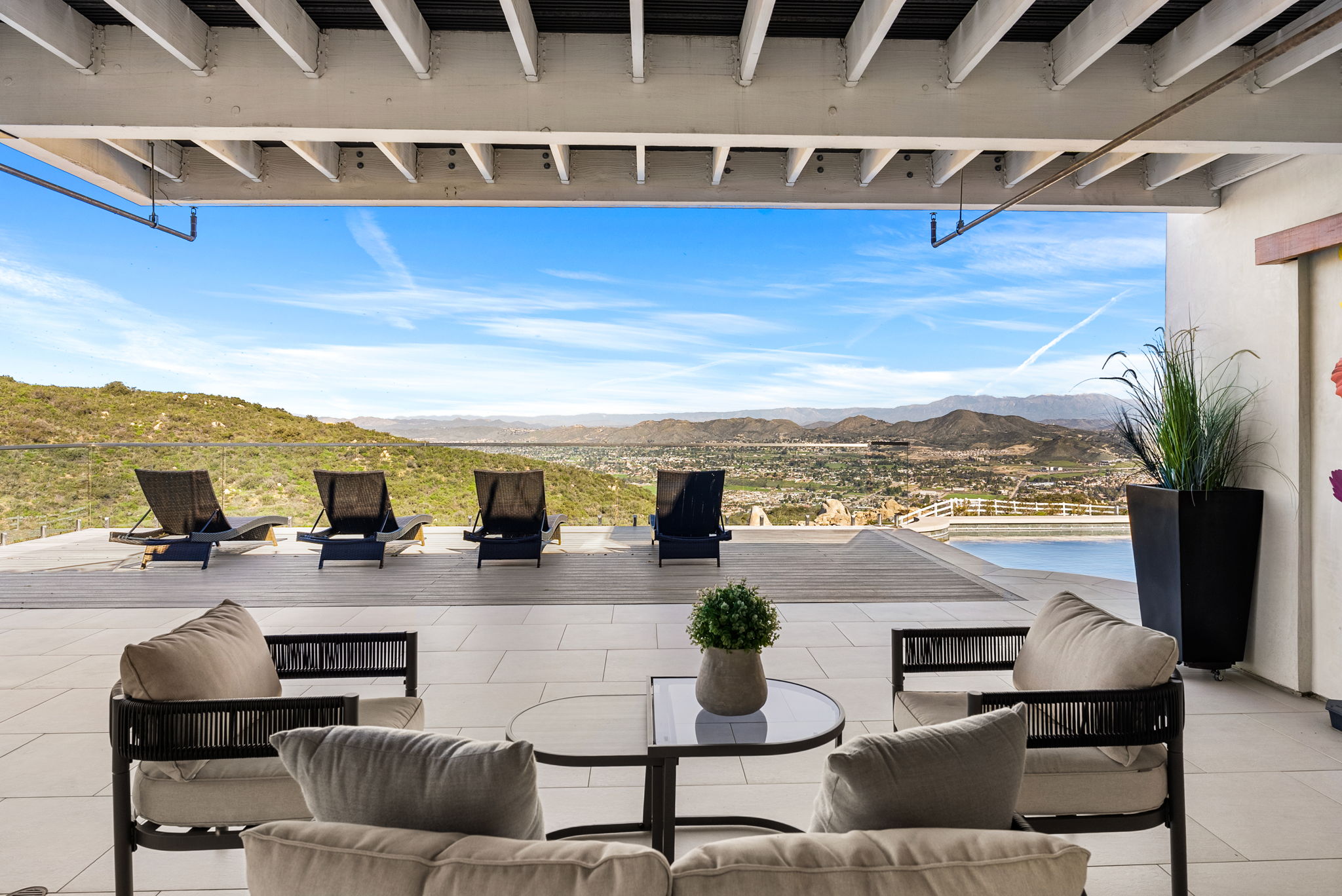 This expansive covered patio offers a luxurious outdoor living space with a commanding panoramic view of the valley and mountains beyond. The area is tastefully furnished with modern seating, including lounge chairs overlooking a sleek infinity-edge pool, creating a perfect environment for relaxation and entertaining. The sophisticated architectural design, featuring clean lines and high-end materials, seamlessly blends indoor comfort with the breathtaking natural landscape.