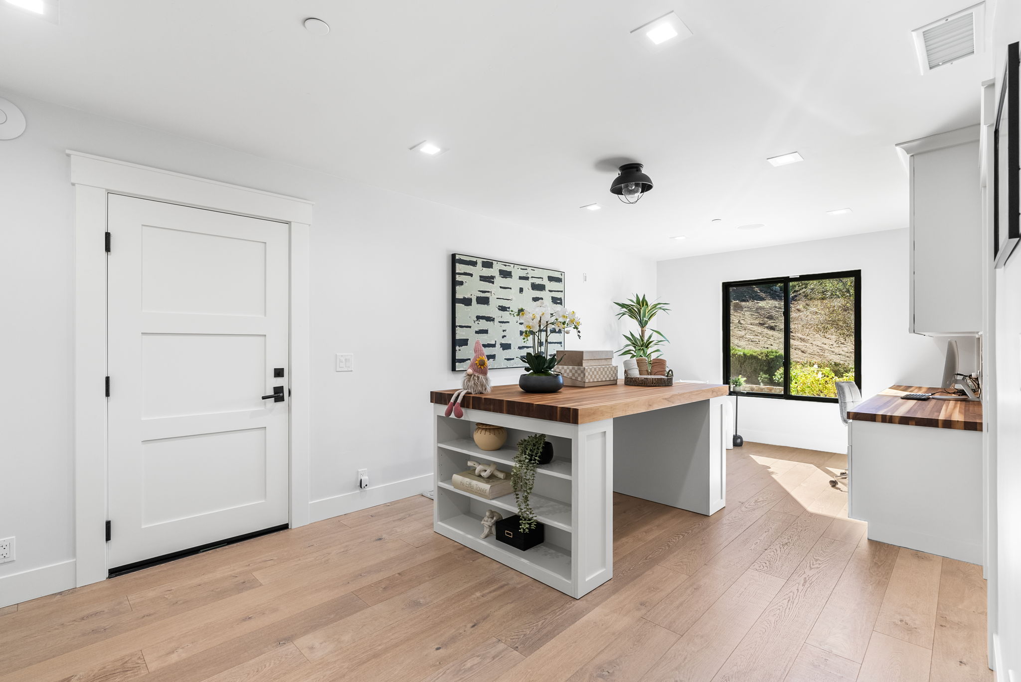 This modern, light-filled home office features wide-plank wood flooring and crisp white walls, creating a clean and spacious atmosphere. The room is anchored by a large, functional island with built-in shelving, complemented by an additional desk surface along the window wall that provides an inspiring outdoor view. Minimalist design choices and contemporary lighting fixtures give the space a professional yet inviting feel.