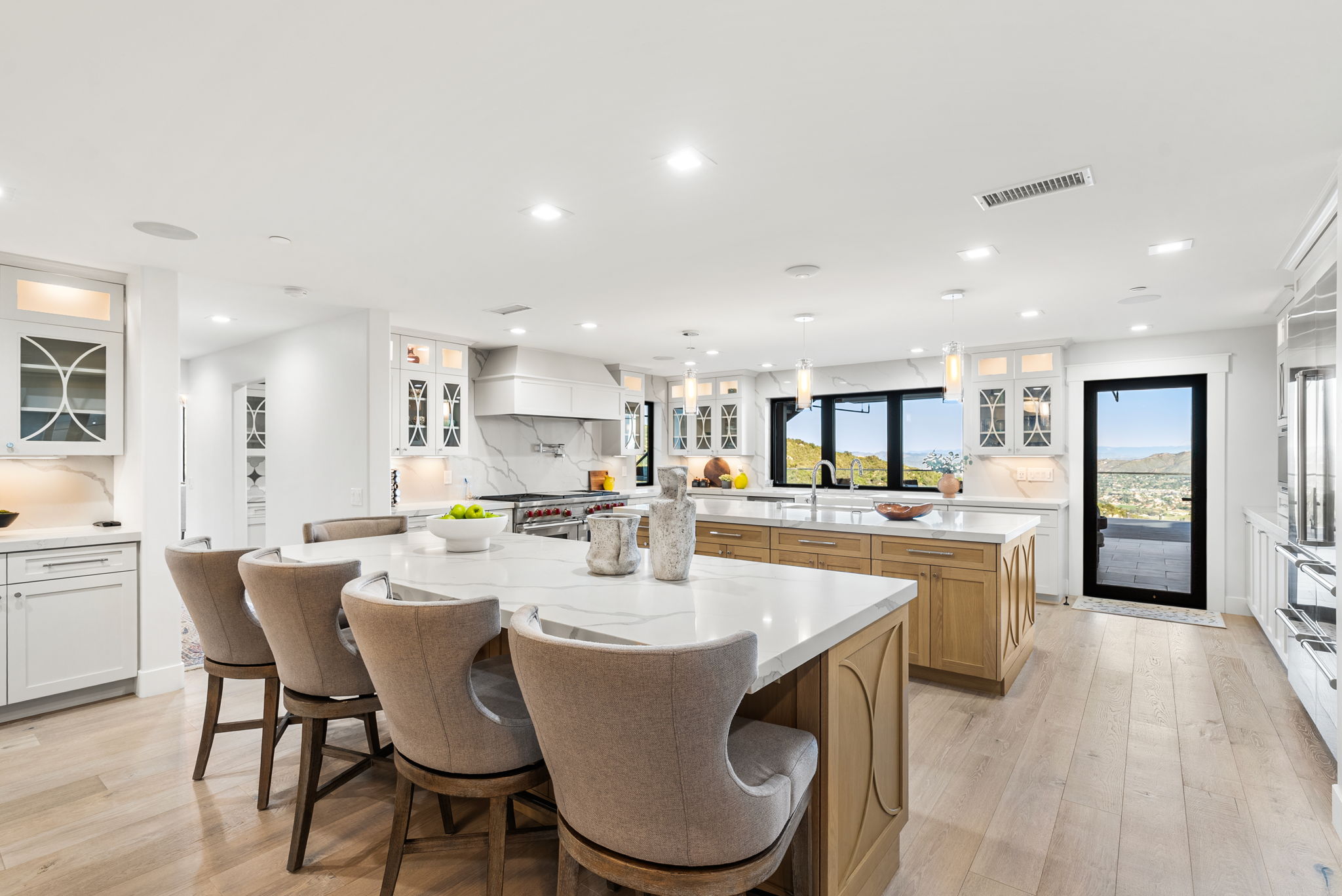 This stunning contemporary kitchen features a spacious dual-island layout with light-toned wooden cabinetry, white marble-style quartz countertops, and a full suite of professional-grade stainless steel appliances. The space is bright and airy, highlighted by recessed lighting, chic pendant fixtures, and a large window overlooking scenic hillside views. Elegant upholstered bar stools add a touch of comfort and style to the expansive island, creating a perfect environment for both cooking and casual dining.
