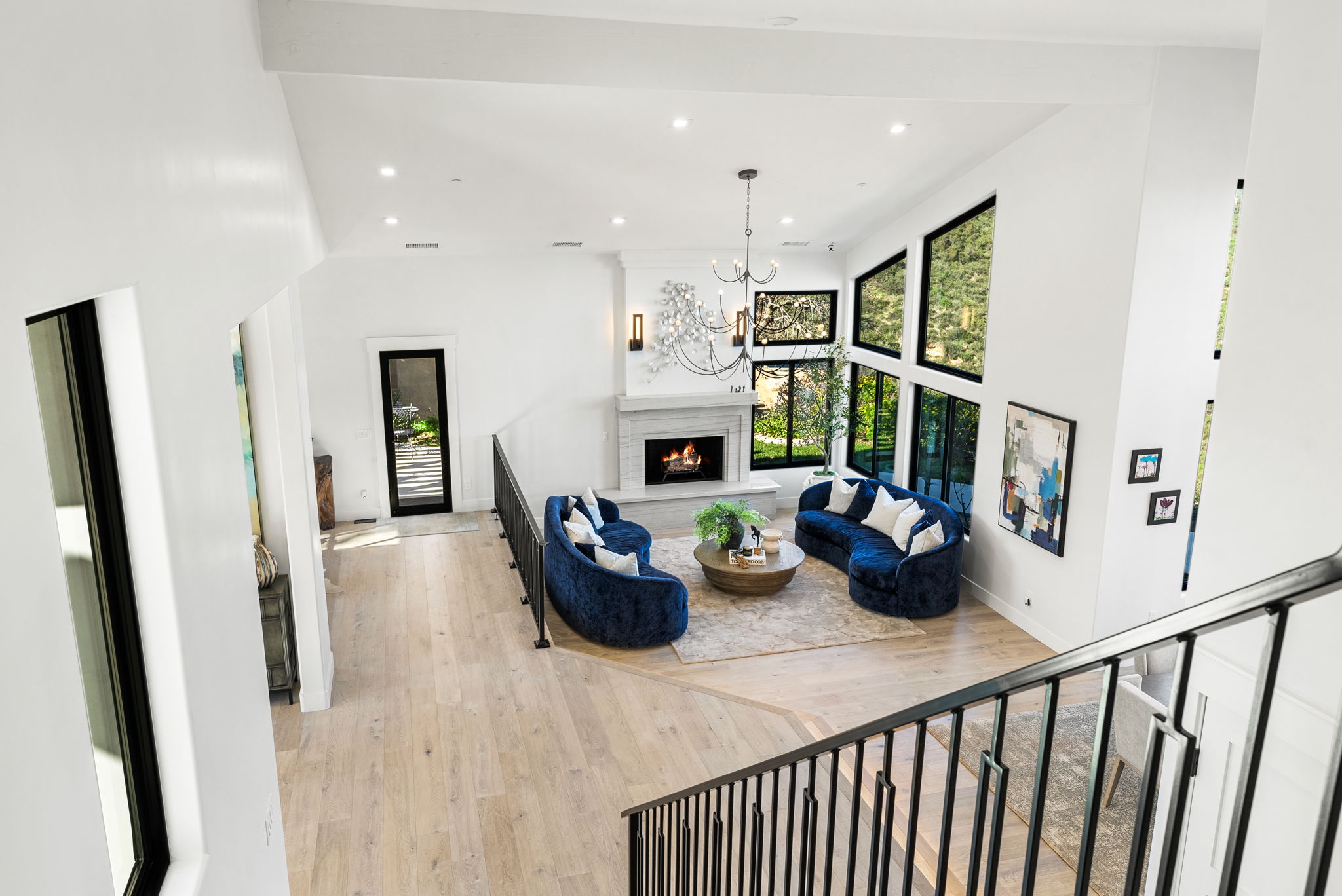 This spacious, modern living room is characterized by a high, vaulted ceiling and an open-concept layout that flows from an upper-level viewpoint. The space is anchored by a sophisticated fireplace, flanked by dual velvet-upholstered, curved blue sofas that create a cozy conversational area. Large, multi-pane windows flood the room with natural light, offering views of the surrounding greenery and enhancing the airy, elegant atmosphere.